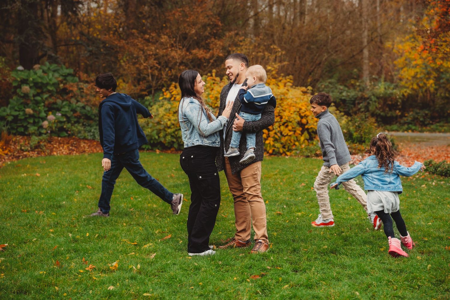 Family of five enjoying autumn park, parents holding toddler while kids play on green grass with fall foliage backdrop.