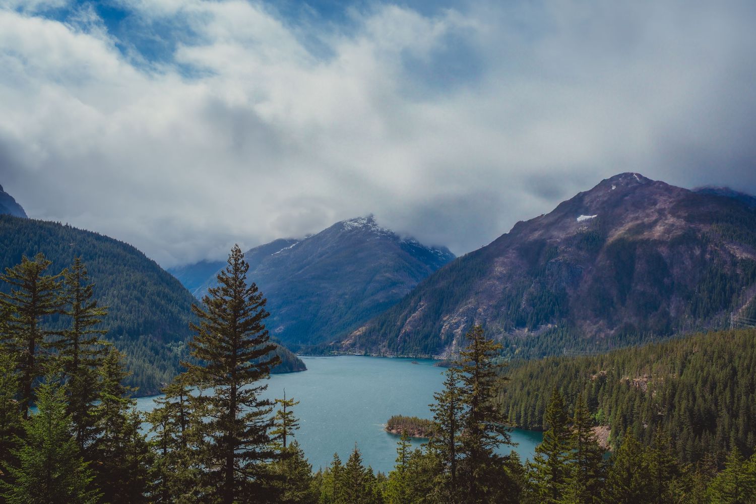 Scenic turquoise mountain lake surrounded by evergreen trees and misty peaks under a cloudy sky.