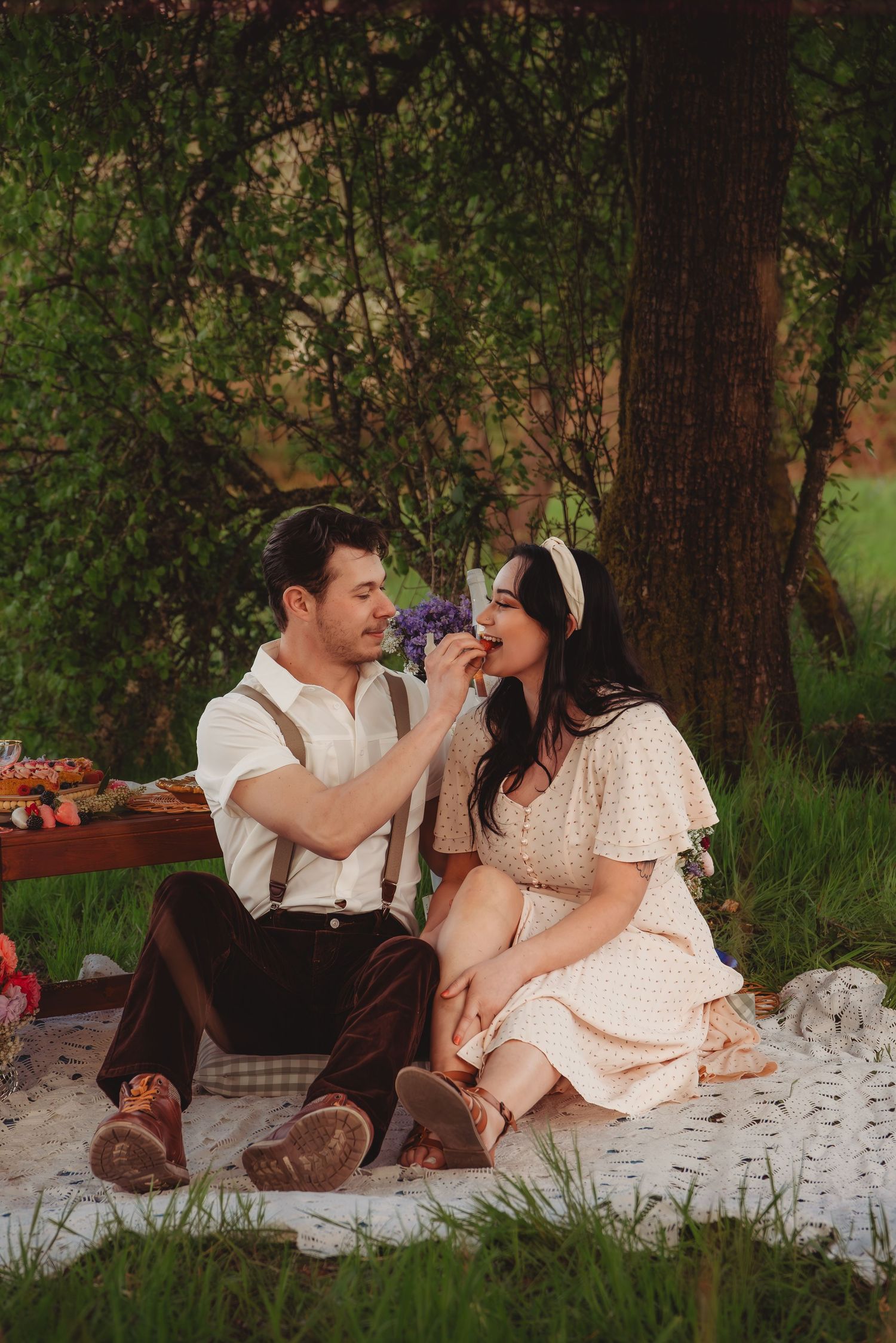 A couple enjoys a romantic picnic under a tree, man in suspenders offers flowers to woman in vintage dress.