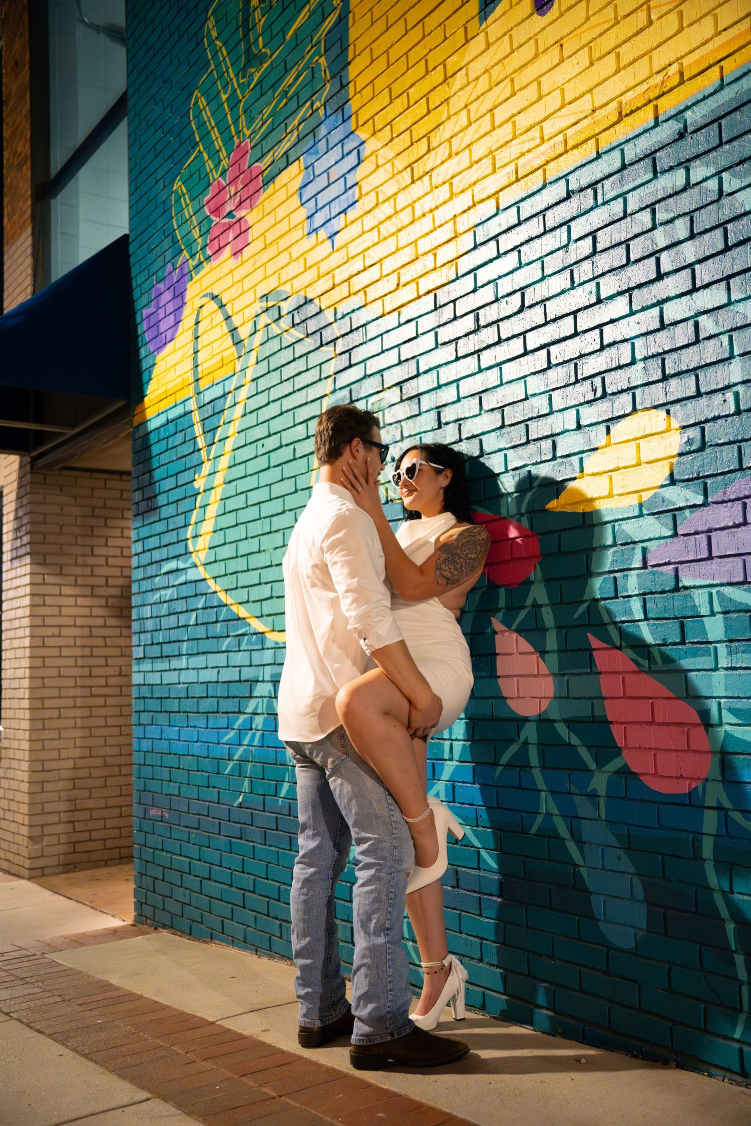 Couple kissing against a vibrant teal and yellow floral brick wall mural on an urban sidewalk.