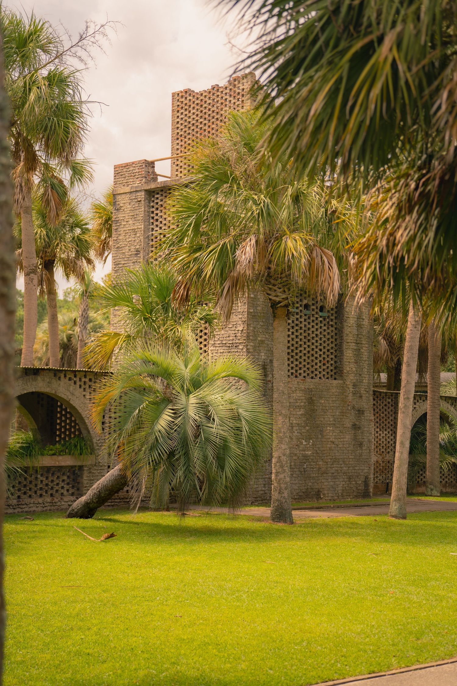 Historic brick tower with decorative lattice walls surrounded by lush palm trees and green lawn under cloudy skies.