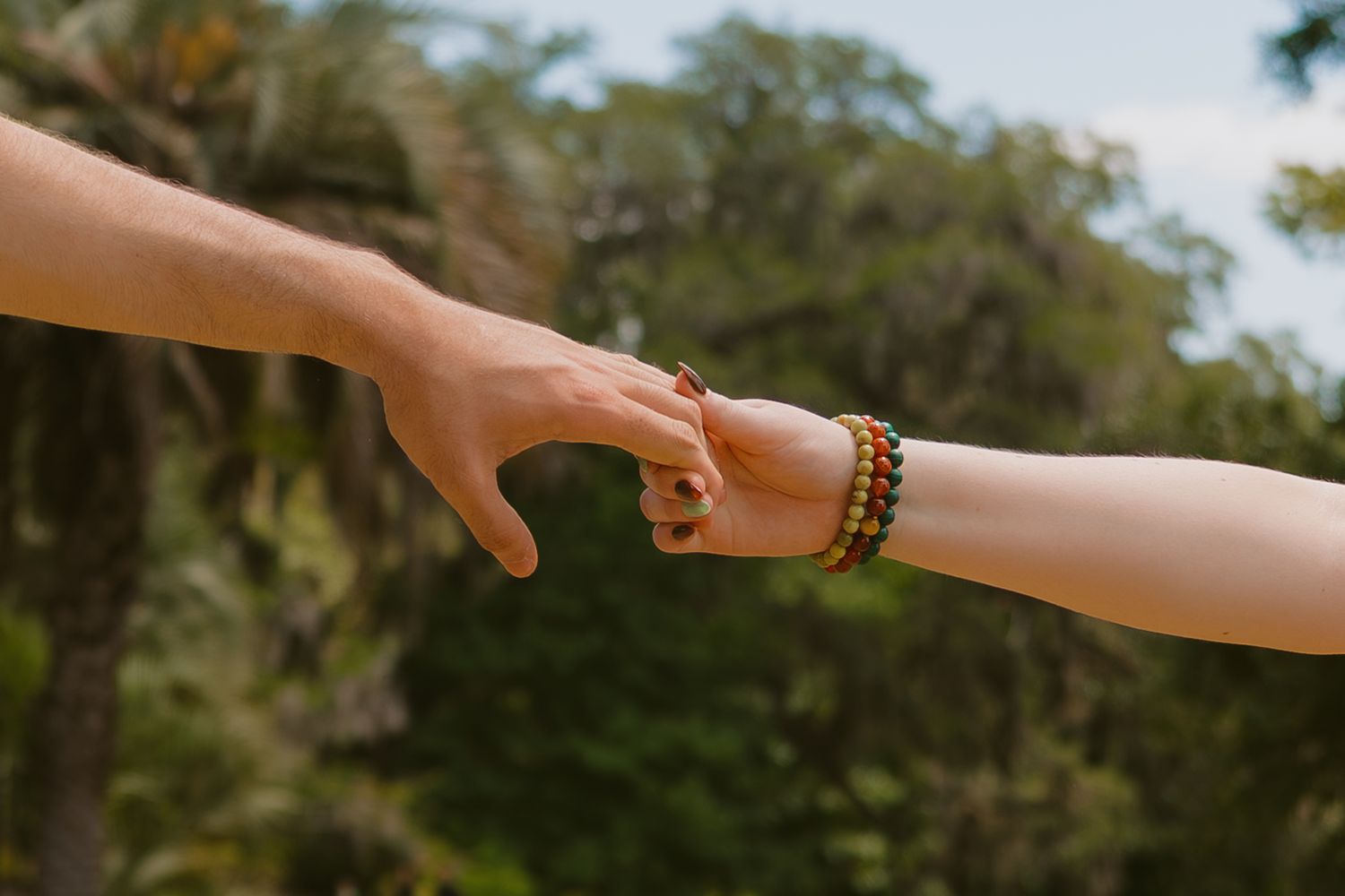 Two hands reaching toward each other outdoors, one wearing beaded bracelets, with lush green trees in the background.
