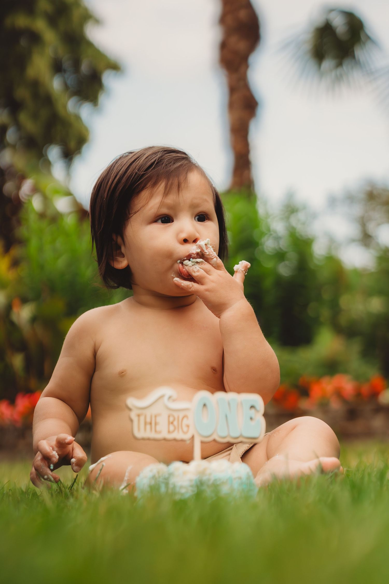 Baby boy enjoying his first birthday cake smash outdoors on green grass with a The Big ONE cake topper.
