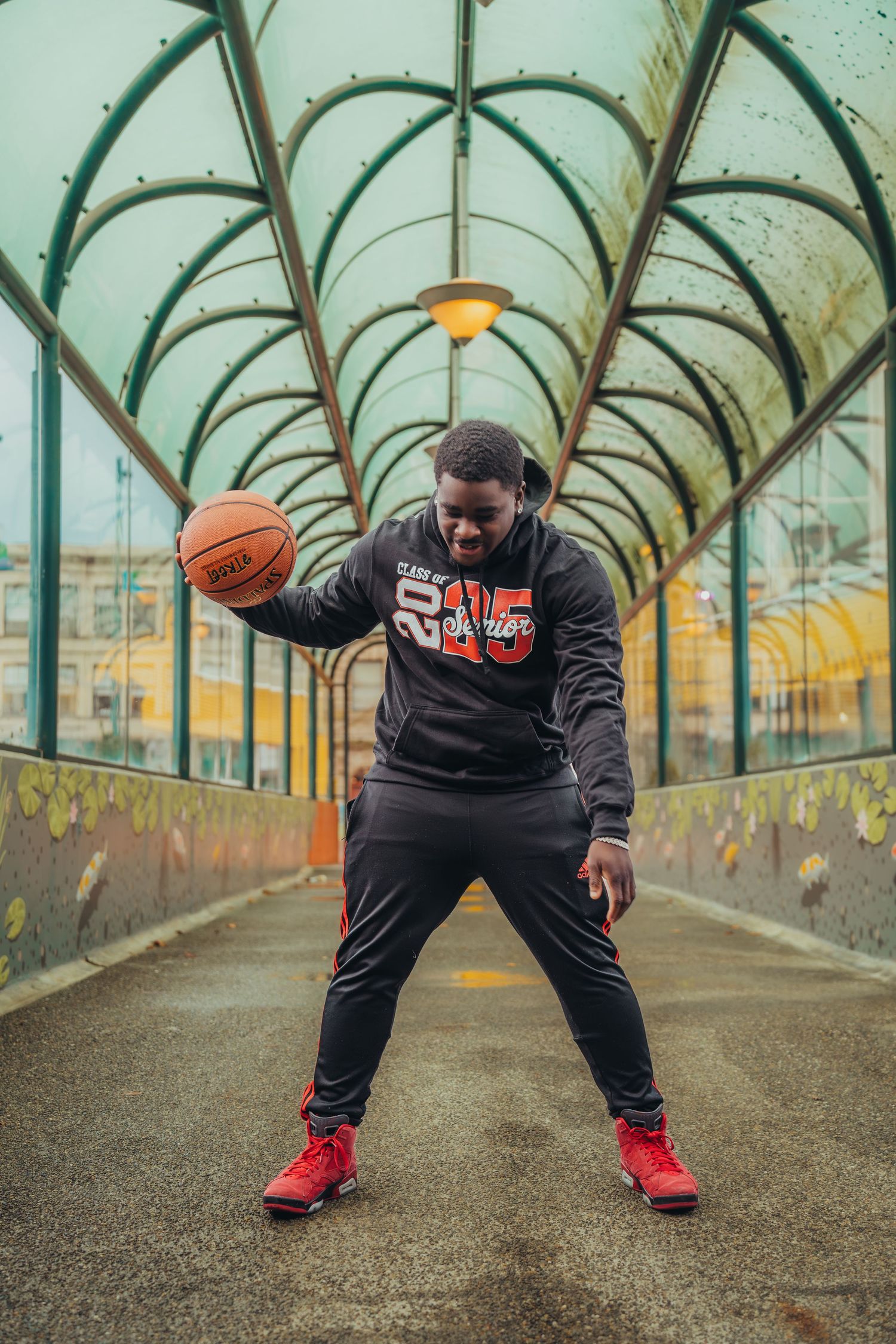 A man in a black hoodie and red sneakers dribbles a basketball on a covered bridge walkway.