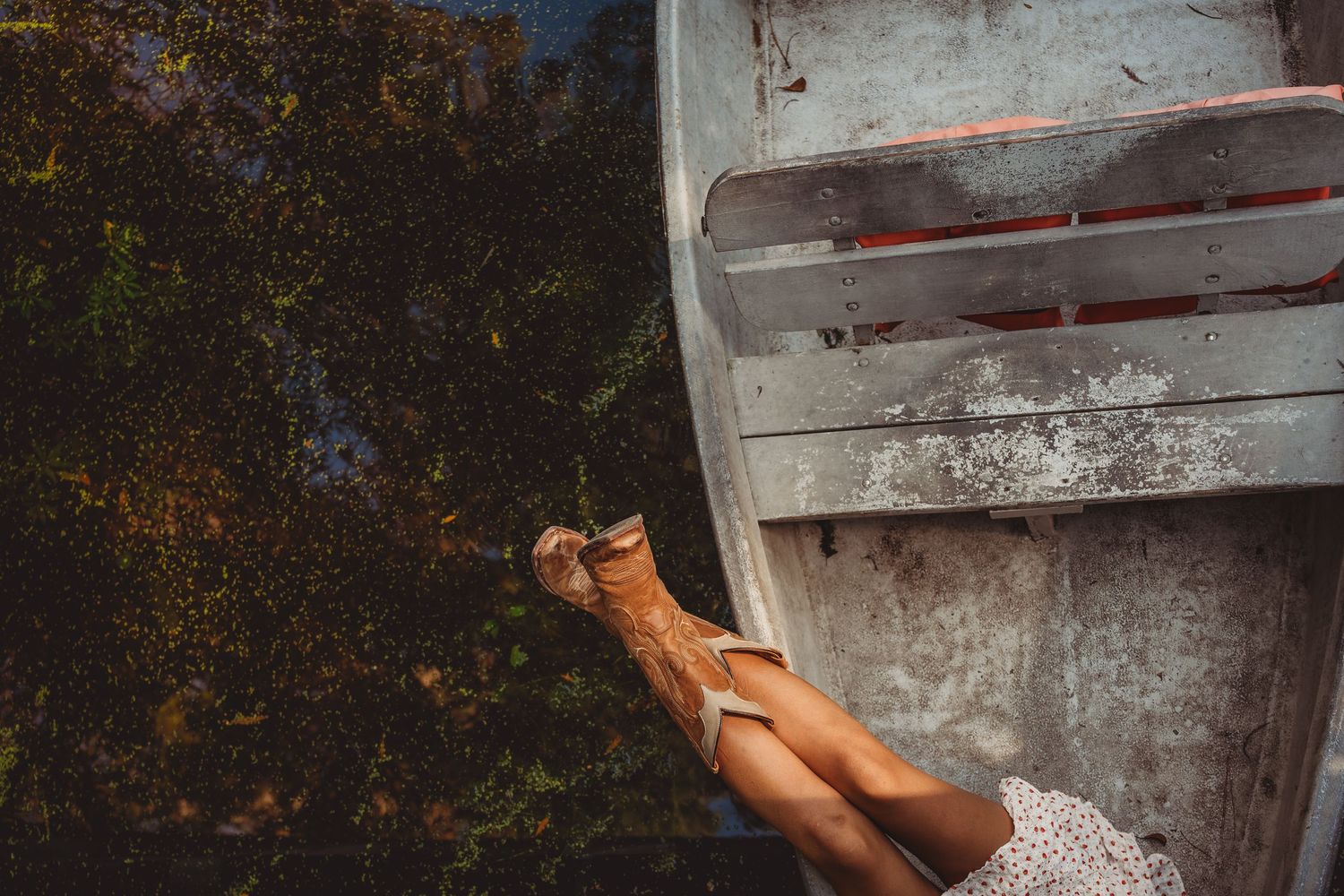 Woman in a floral skirt and sandals relaxing beside a weathered aluminum rowboat near a tranquil pond.