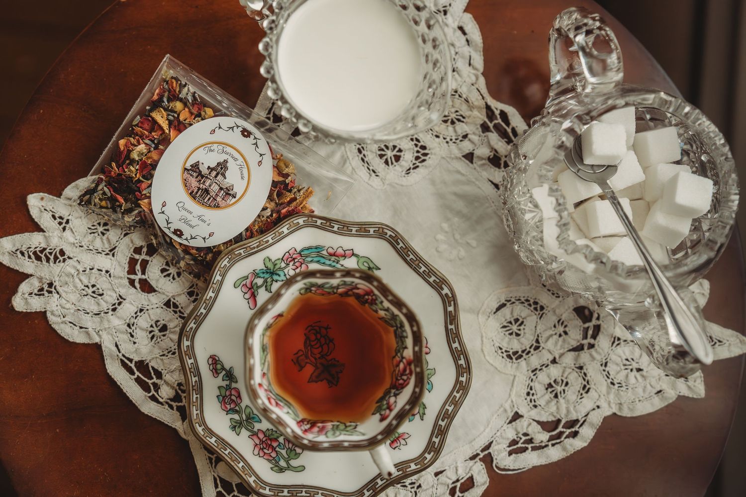Elegant china teacup with brewed tea, sugar cubes, milk, and lace doily on antique wooden table.