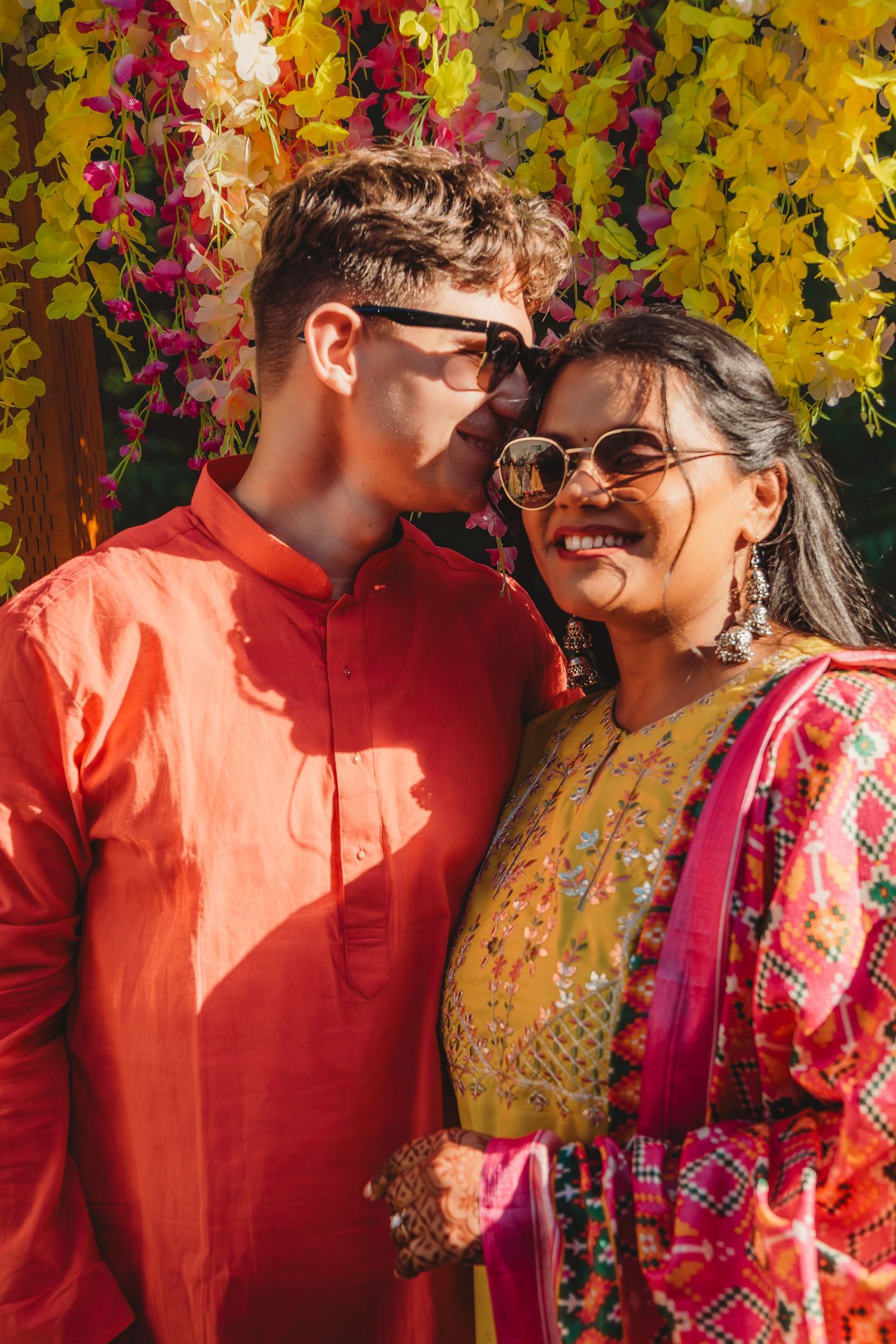 Couple in traditional Indian attire smiling together under colorful floral decorations at a festive celebration.