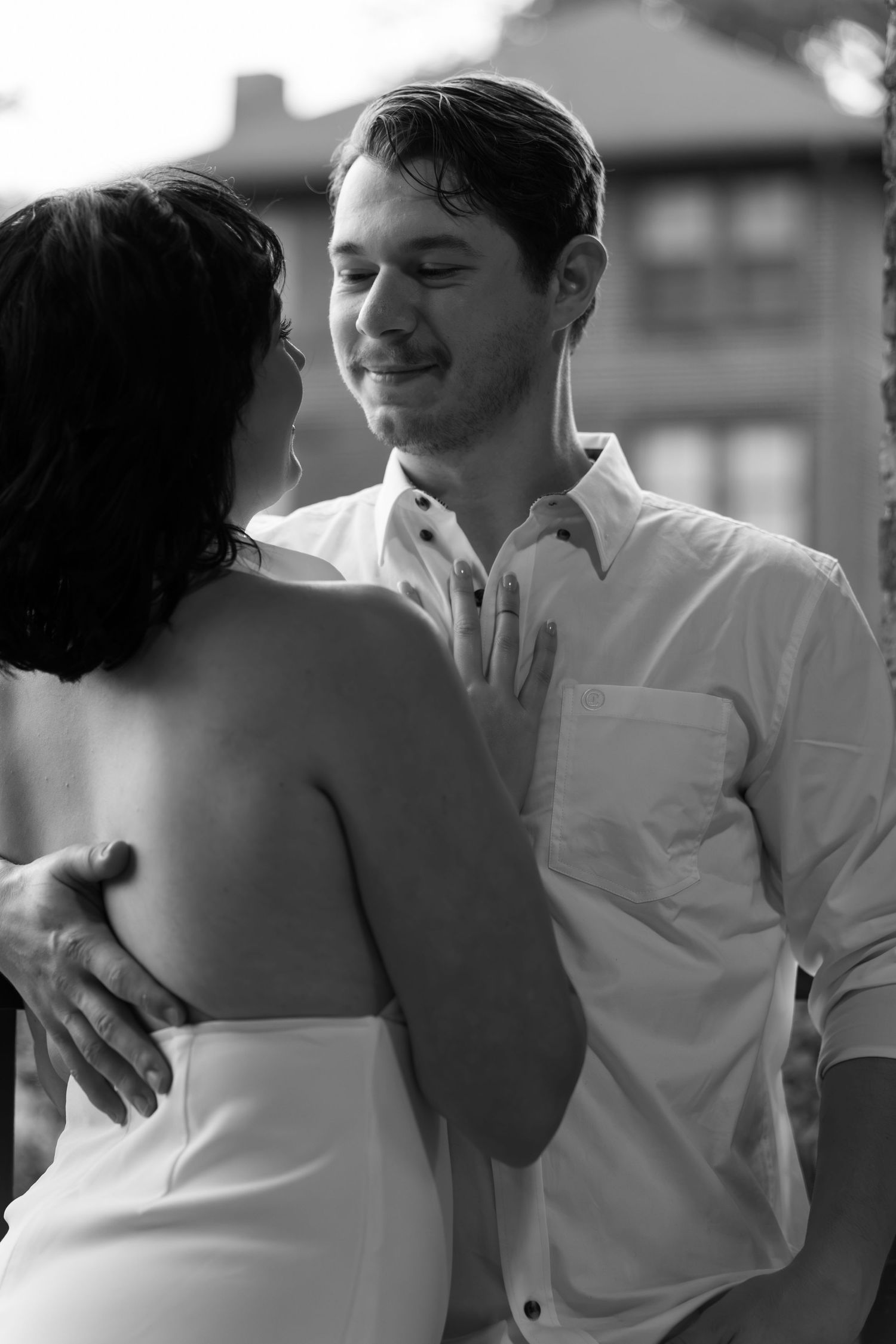 Black and white portrait of a smiling man in a white button-up shirt embracing a dark-haired woman in a white dress outdoors.