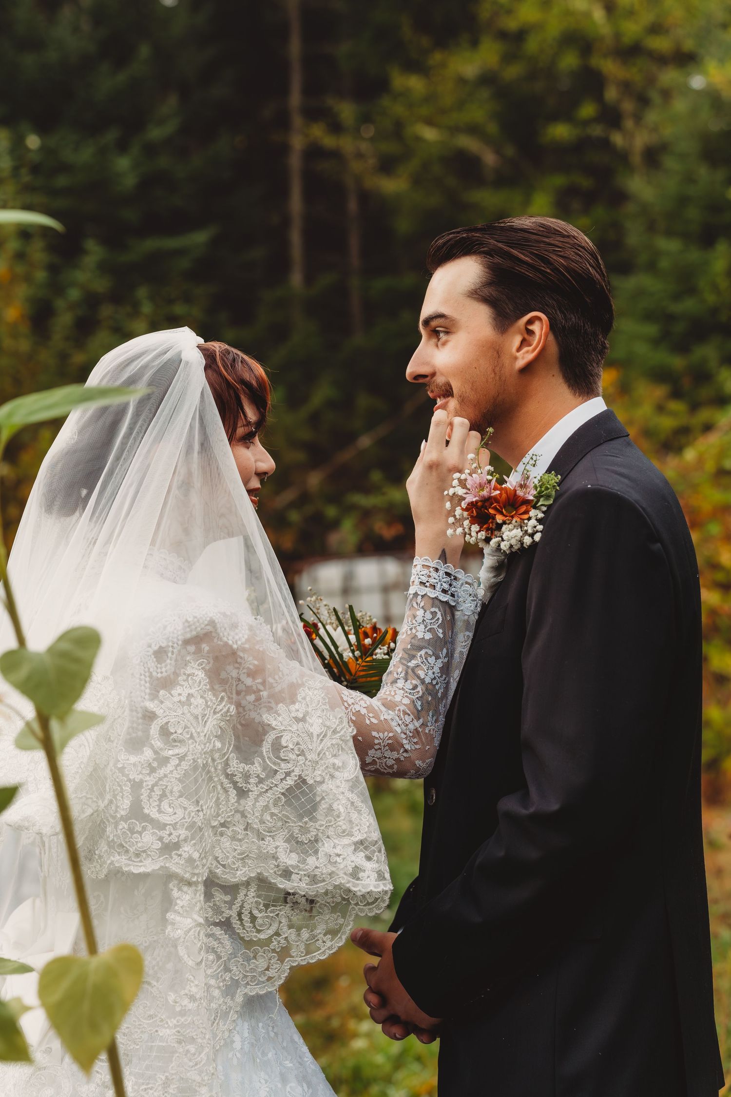 Bride in lace gown and veil tenderly touches groom's face during outdoor wedding ceremony in forest.