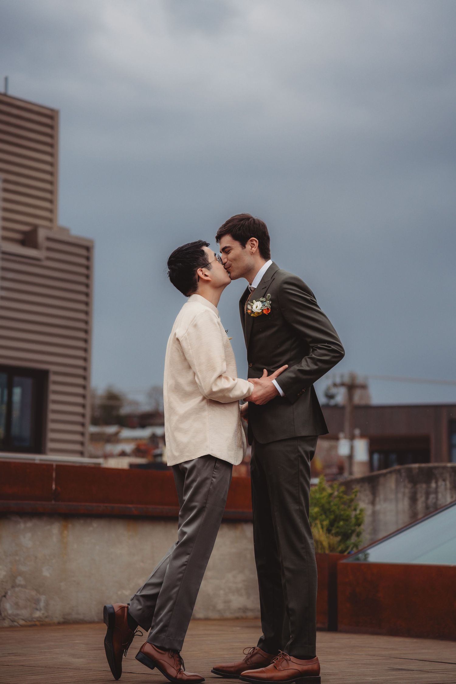 Two men sharing a romantic kiss on a rooftop with a city skyline backdrop on a cloudy day. Seattle