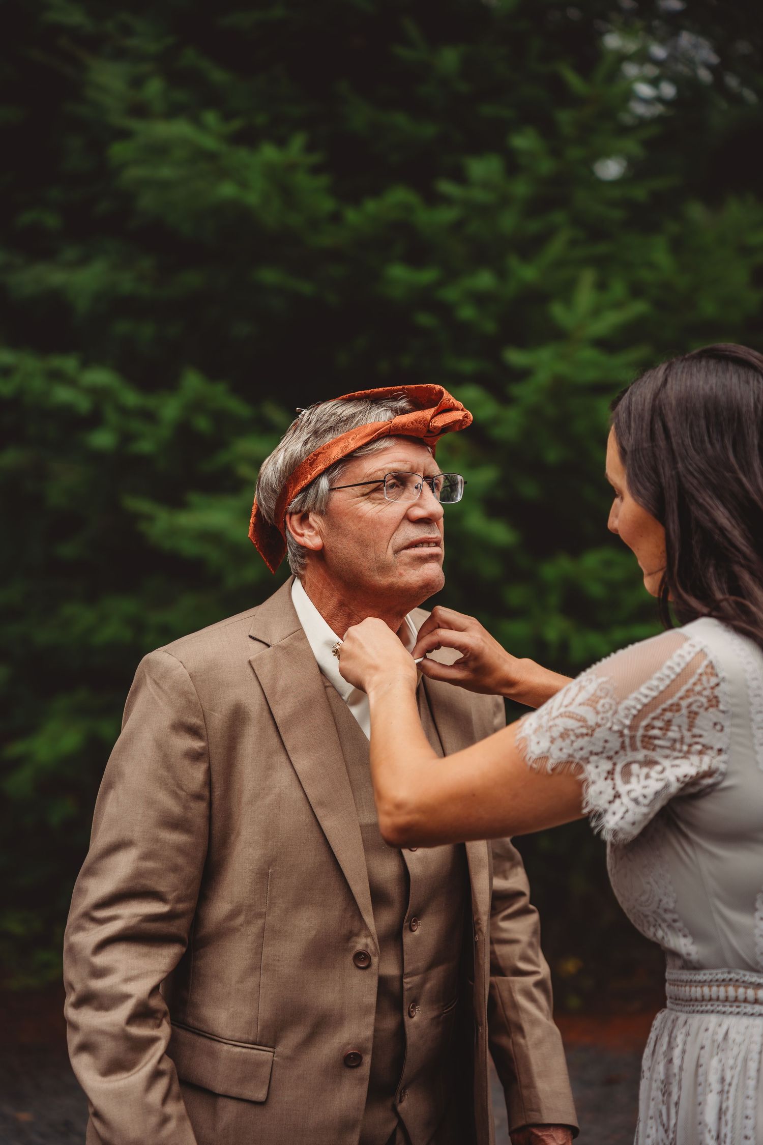 Woman adjusting mans tie outdoors, man wearing orange headband and tan suit with green trees background.