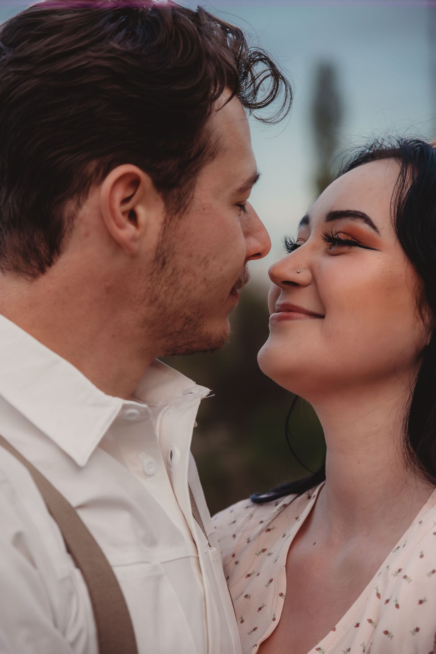 A romantic couple shares an intimate moment, almost kissing, with the woman smiling softly wearing floral dress and orange eye makeup.