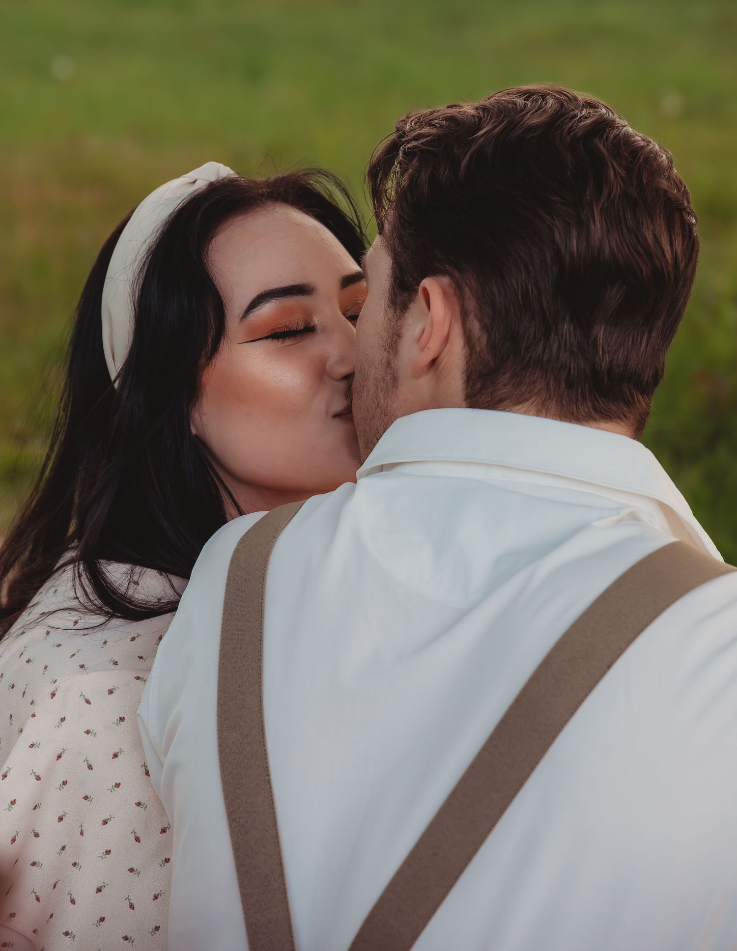 A couple shares a romantic kiss outdoors, the woman wearing a white headband and bold orange eye makeup.