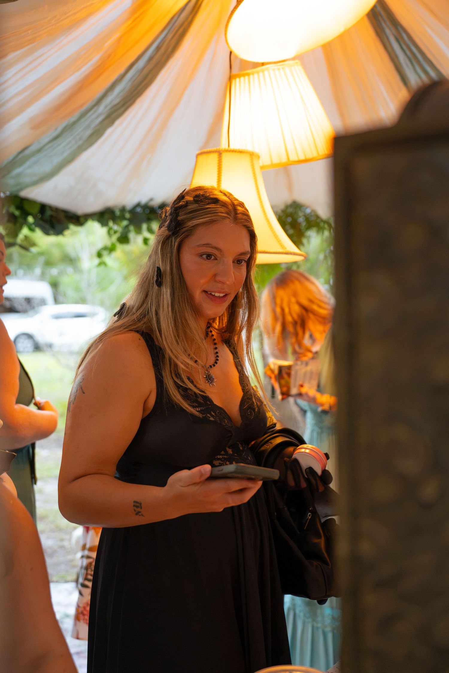 Woman in a black dress holding a phone and dark jacket smiles inside a draped tent with warm lamps at an outdoor event.