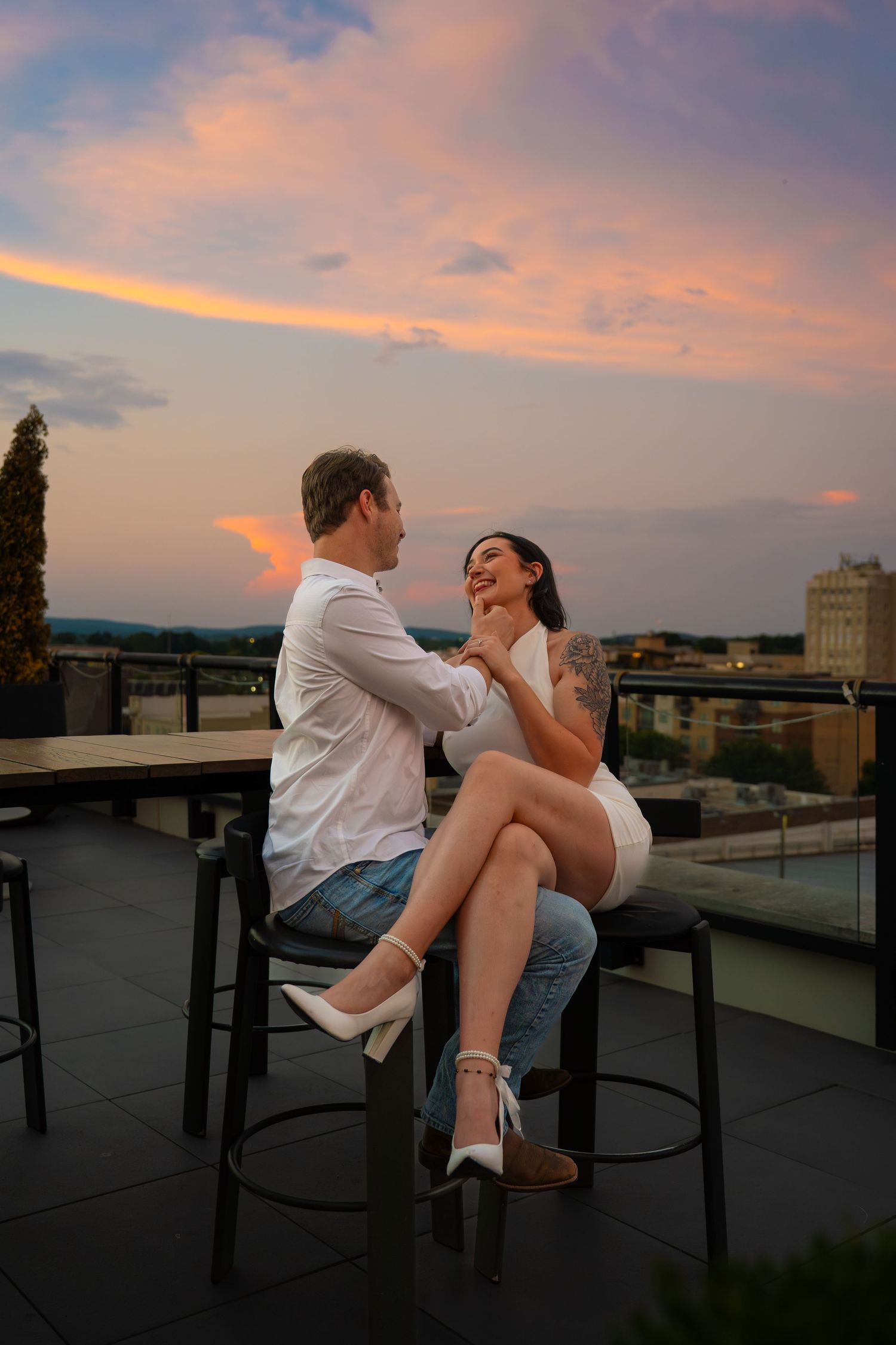 Couple sitting close on bar stools holding hands on a rooftop terrace at sunset with a pink and orange sky over the city.