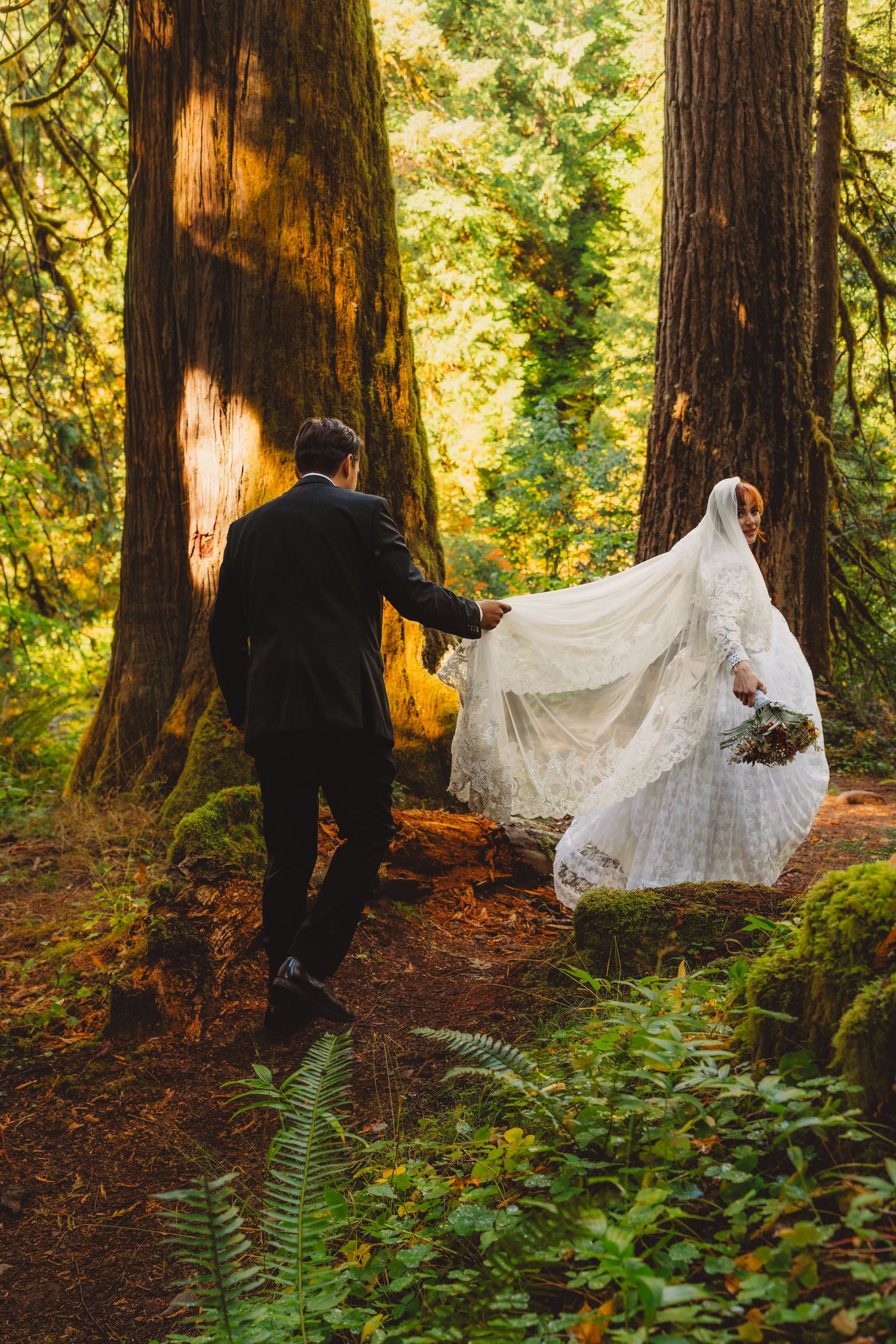 Bride and groom walking through a lush forest, groom holding bride's white lace wedding dress train.