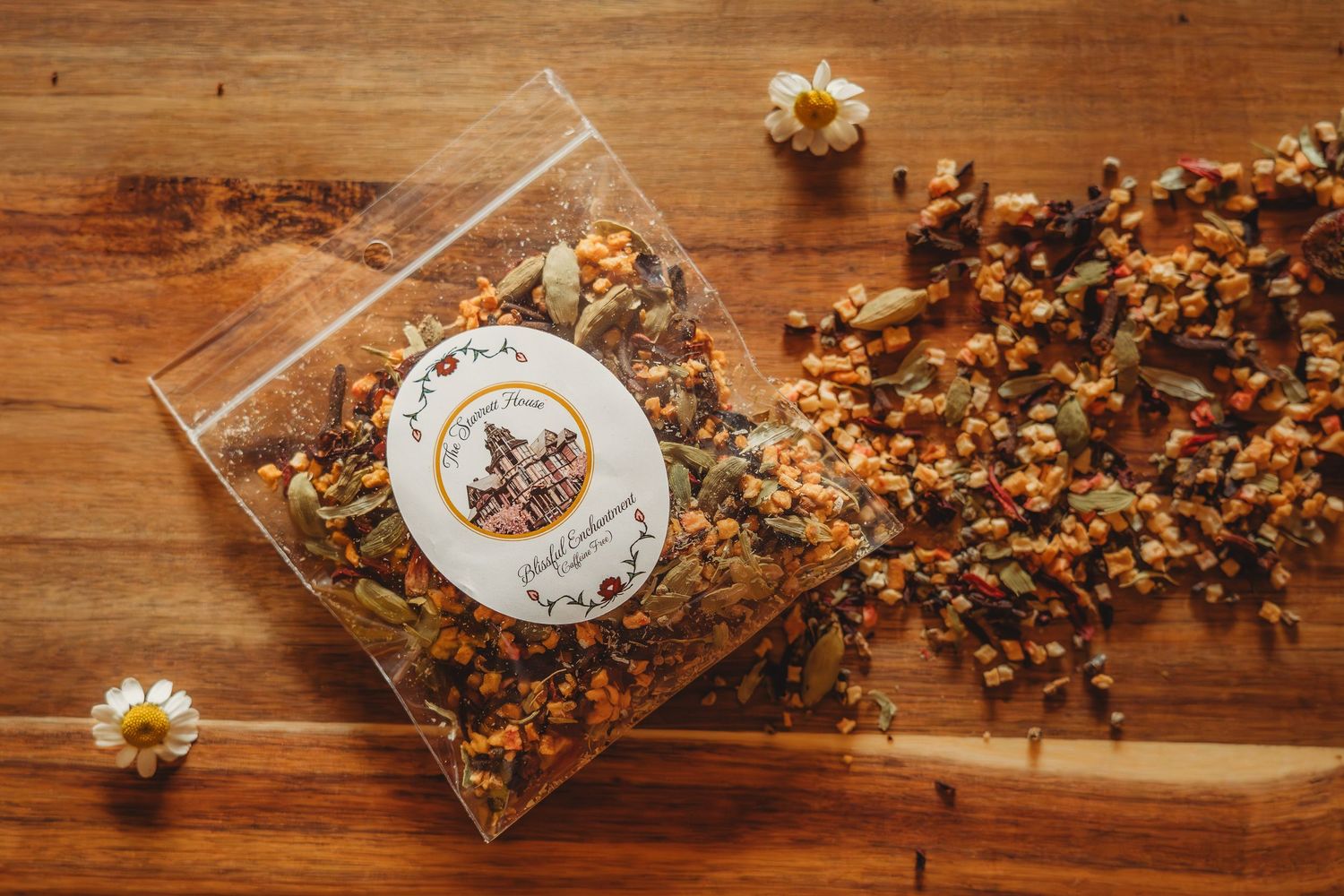 Clear bag of artisan herbal tea blend with floral label on rustic wood surface, surrounded by dried botanicals and daisy flowers.