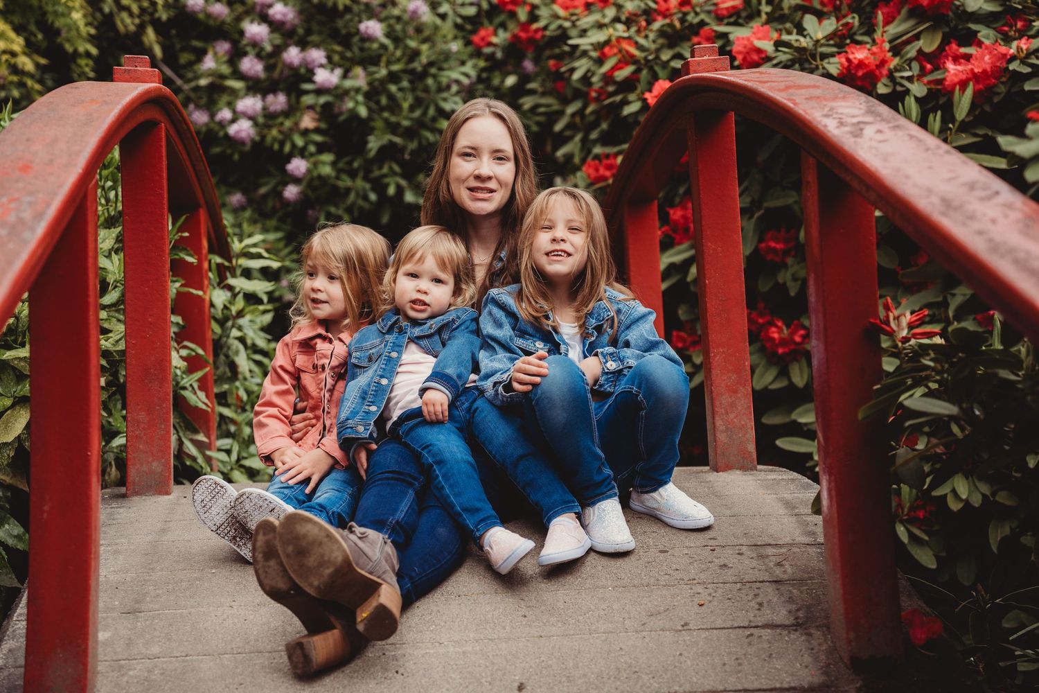 A mother and three young children sitting together on a red garden bridge surrounded by lush greenery and pink flowers.