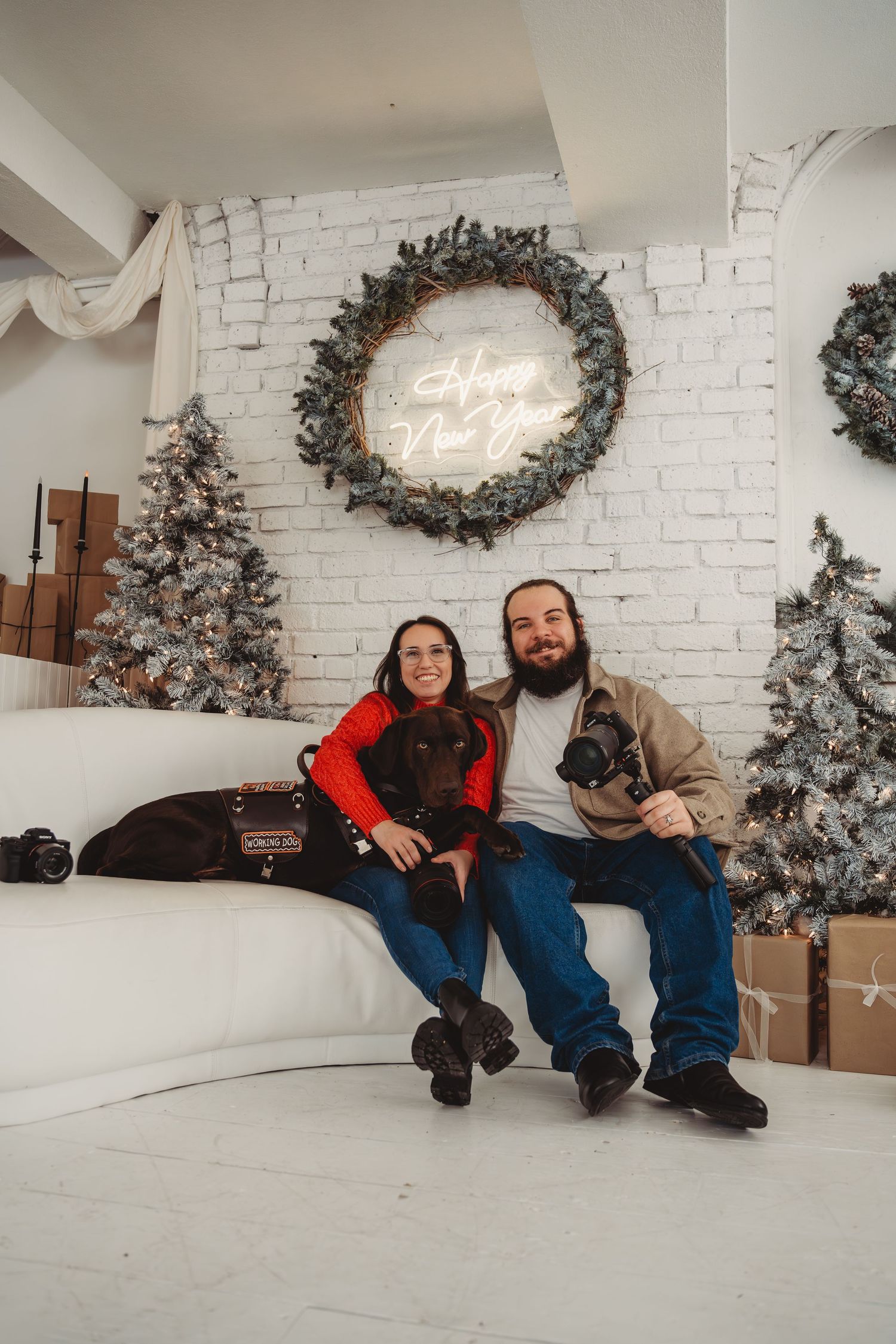 Couple sitting on white sofa with two dogs amid Christmas trees and Happy New Year neon sign decor.