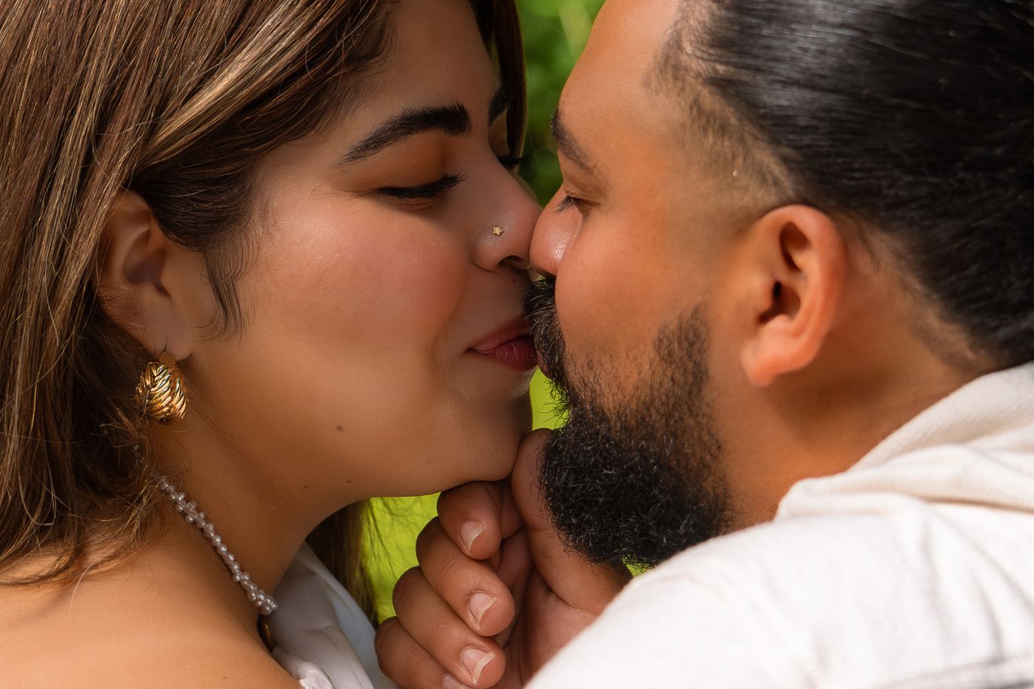 A couple shares a romantic kiss, the woman wearing gold earrings and a pearl necklace, with lush greenery in the background.