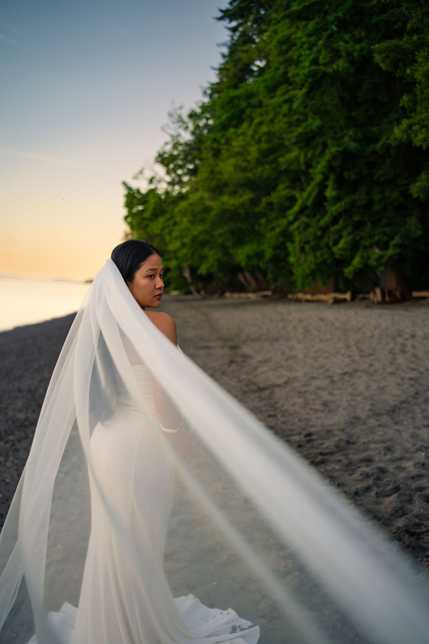 Bride in white wedding dress and flowing veil on sandy beach at sunset with lush green trees.