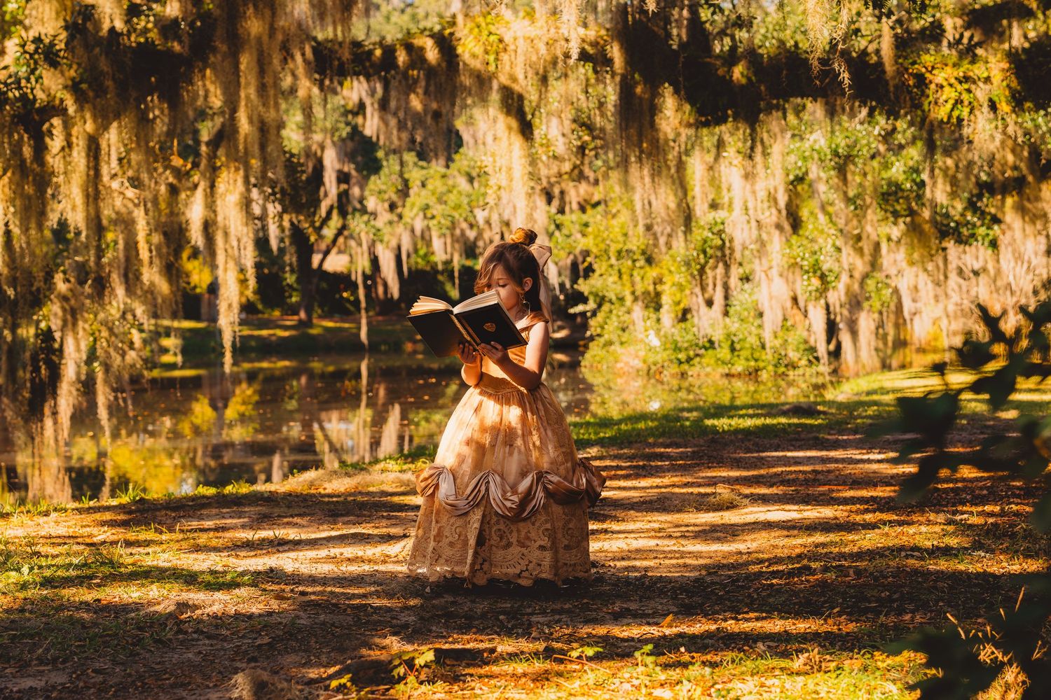 Woman in vintage golden gown reading a book on a sunlit path beneath moss-draped trees.