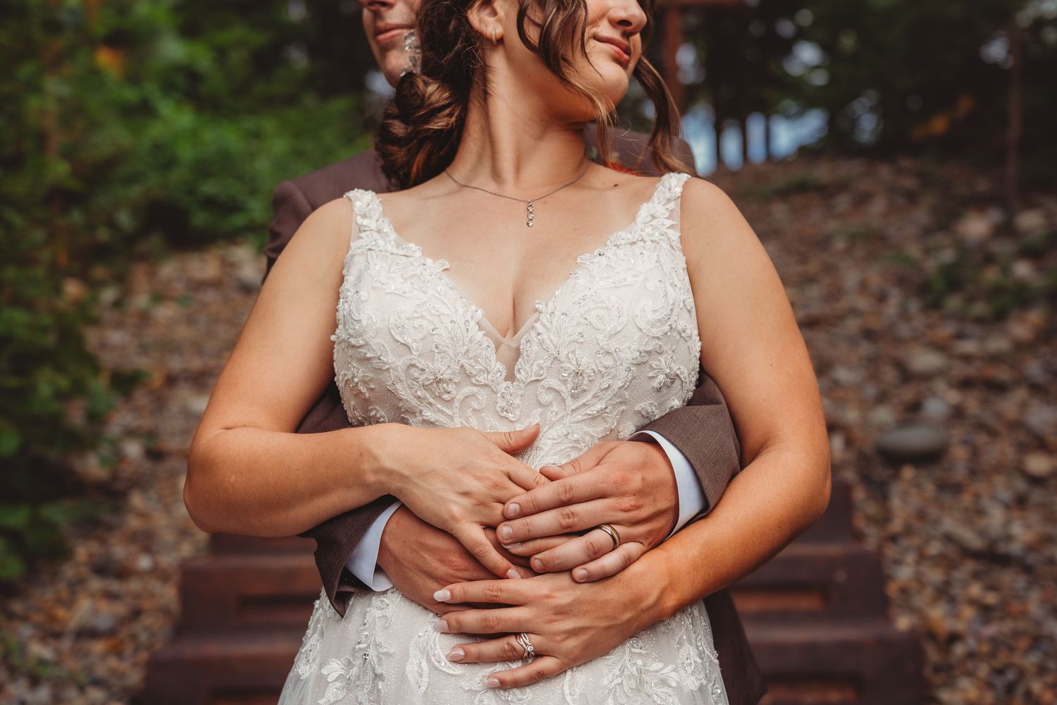 Bride in lace wedding gown embraced by groom in suit, holding her waist outdoors near wooden steps.