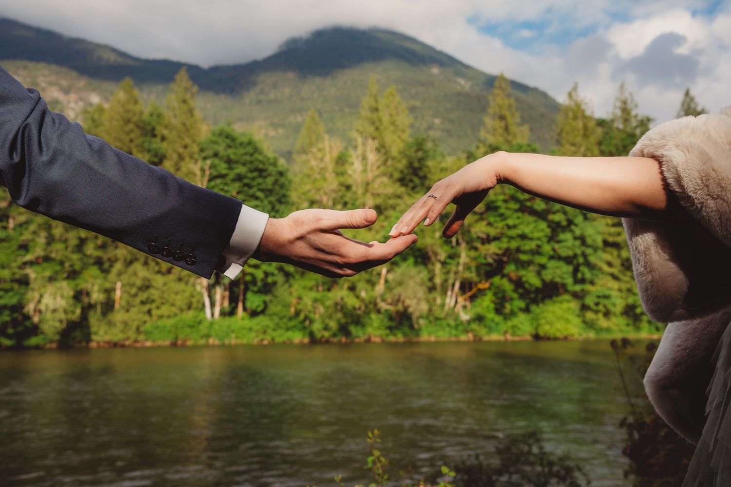 Couple reaching hands toward each other near a river with lush green trees and mountain backdrop.