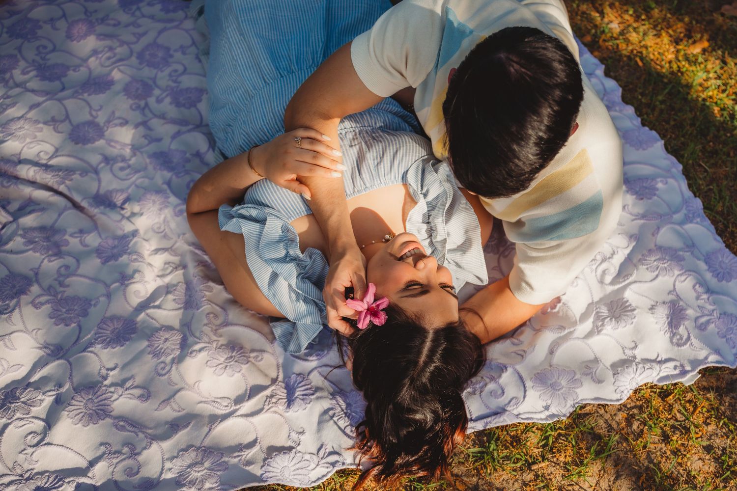 Couple sharing a romantic moment on a white blanket outdoors, woman with pink flower in hair.