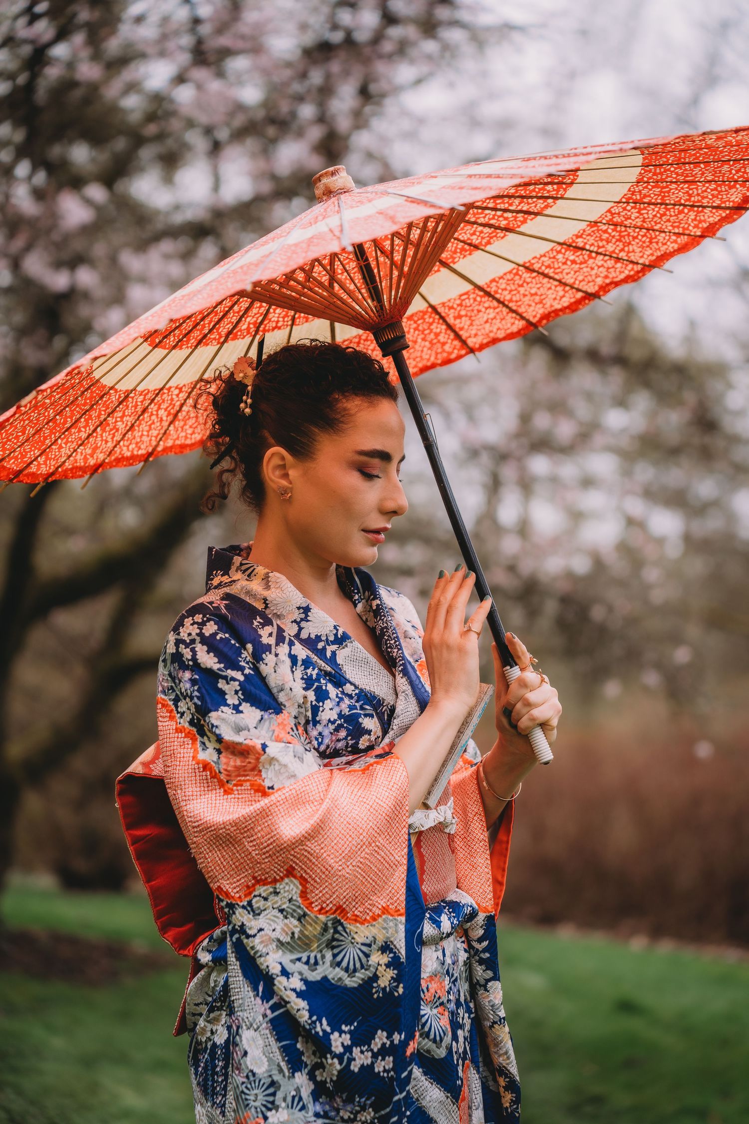 Woman in colorful Japanese kimono holding a red parasol umbrella outdoors near blooming trees.