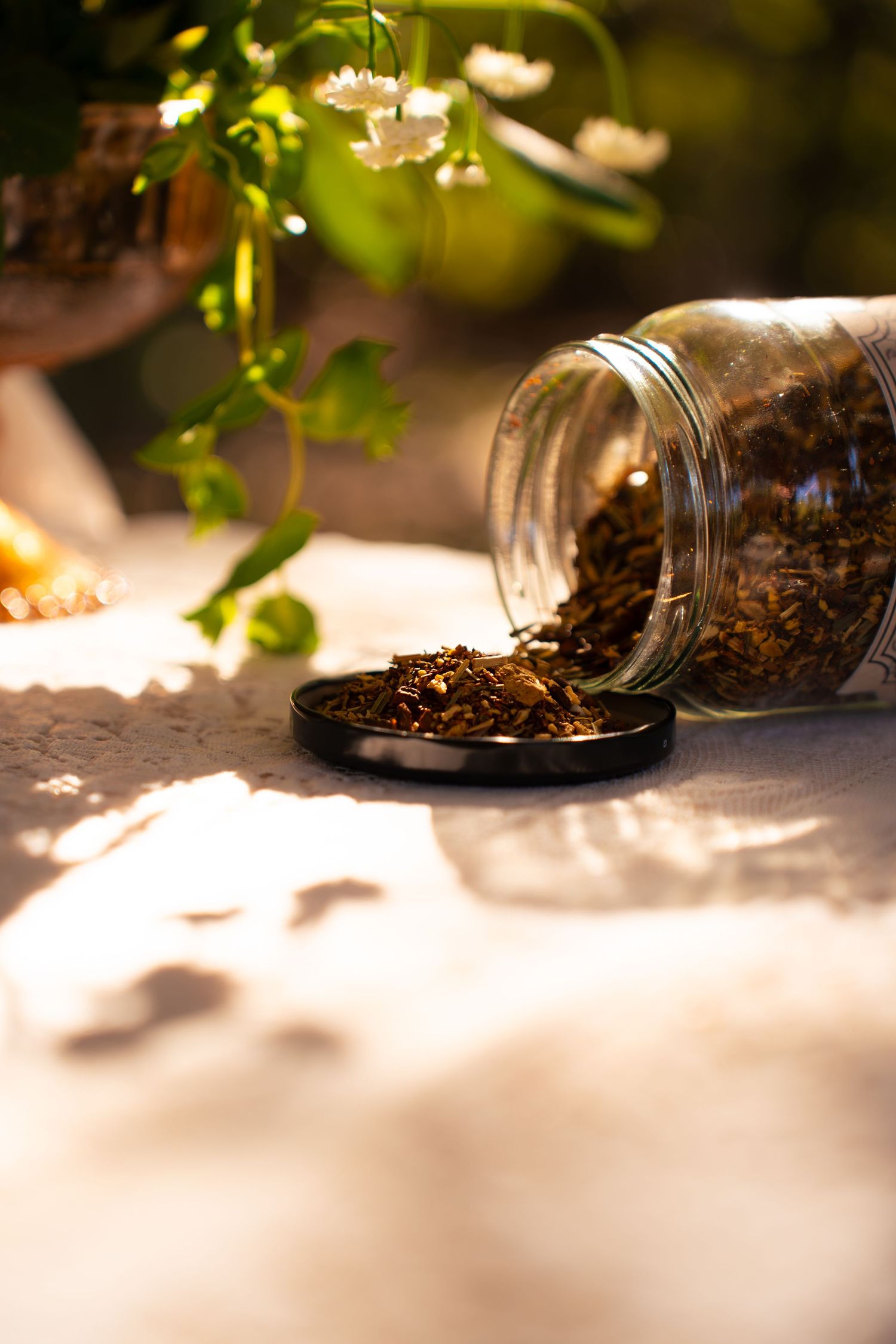 Glass jar of dried loose-leaf herbal tea spilling onto a black lid on a lace tablecloth, with white flowers and green leaves behind.
