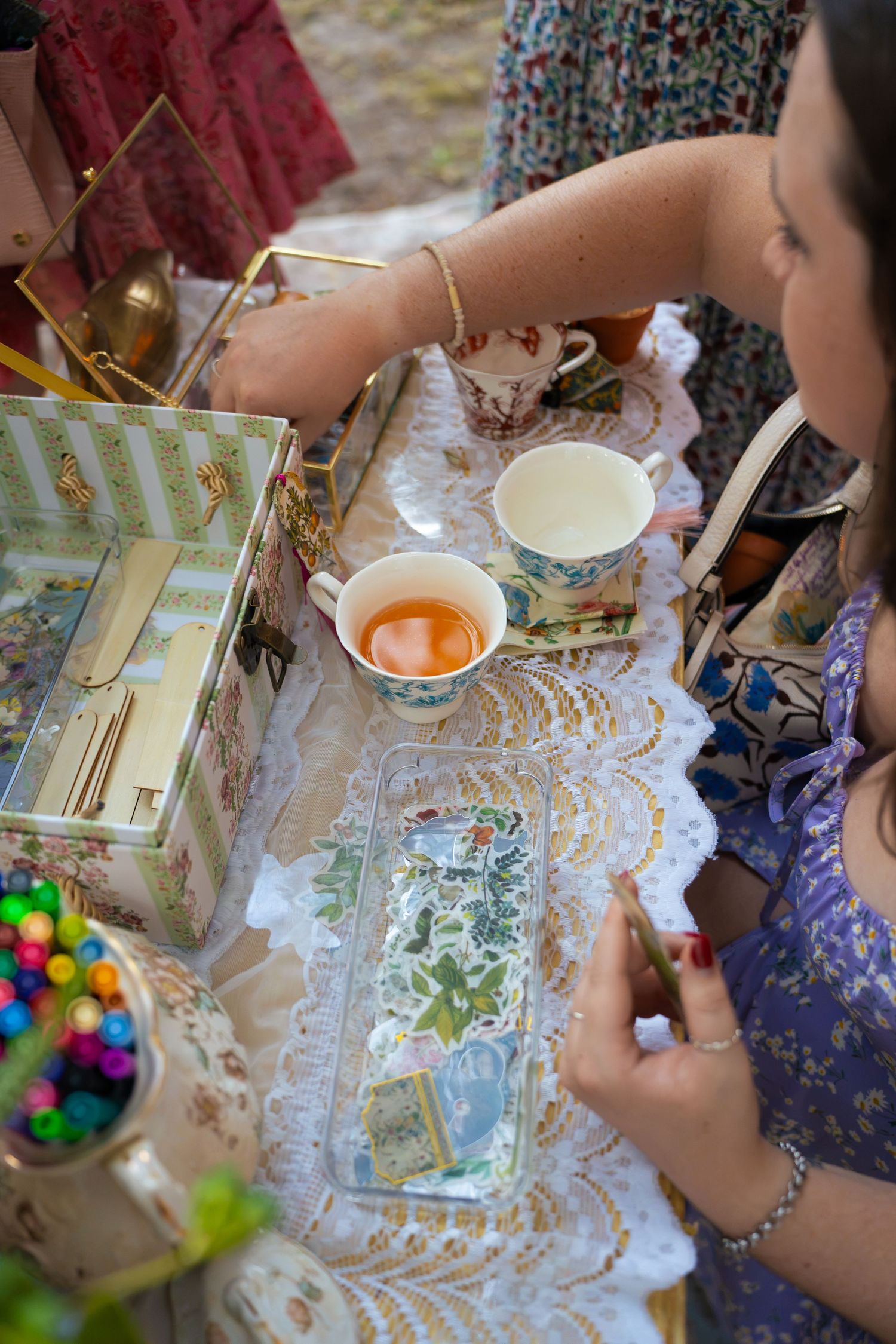Woman in a lavender dress sips tea at a vintage tea party table with lace linens, floral teacups, a decorative box, and colorful candy.