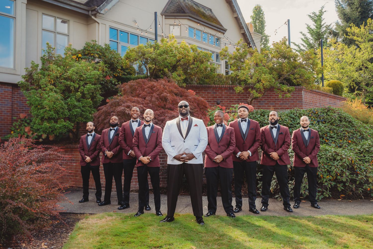 Groom in white tuxedo stands with groomsmen in burgundy suits outside elegant venue.
