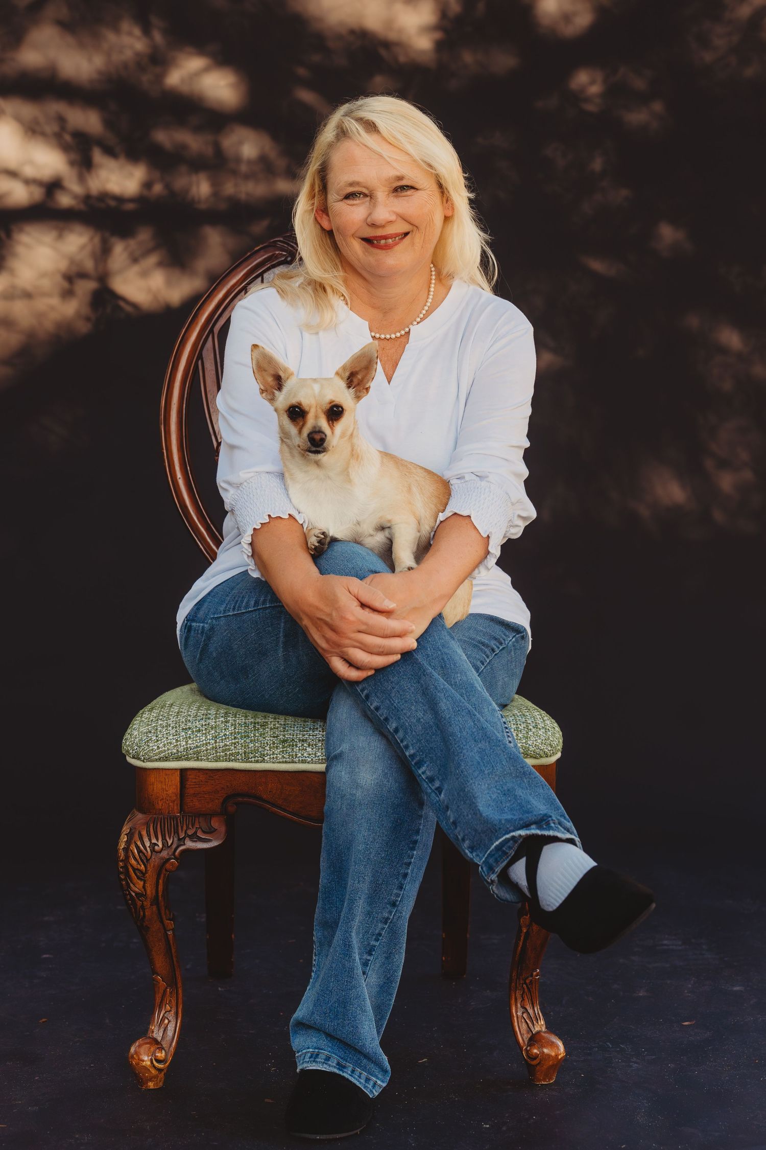Smiling blonde woman sitting on an ornate chair holding a small Chihuahua dog.