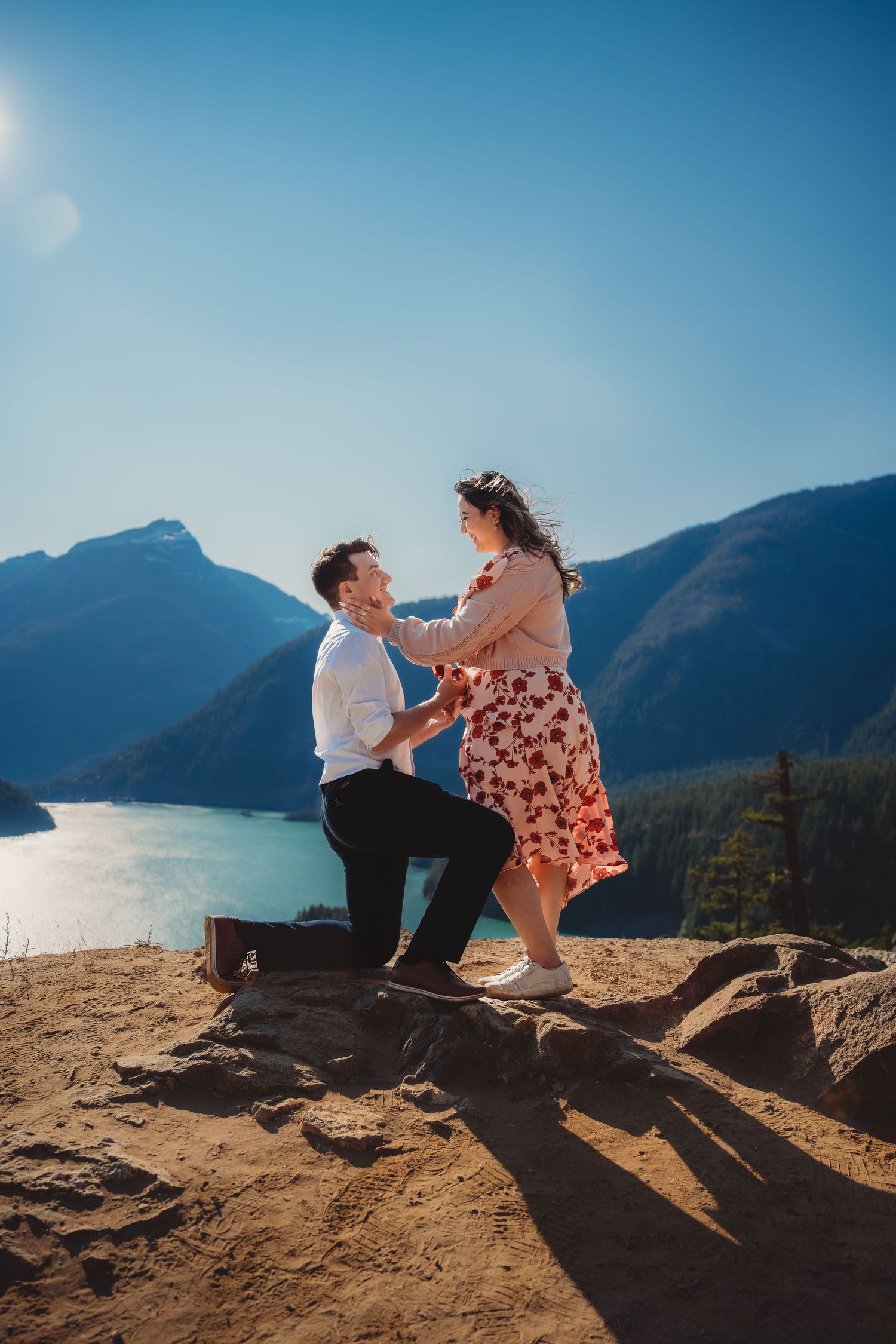 Man proposes on one knee to woman on rocky overlook with mountain lake backdrop under clear blue sky.