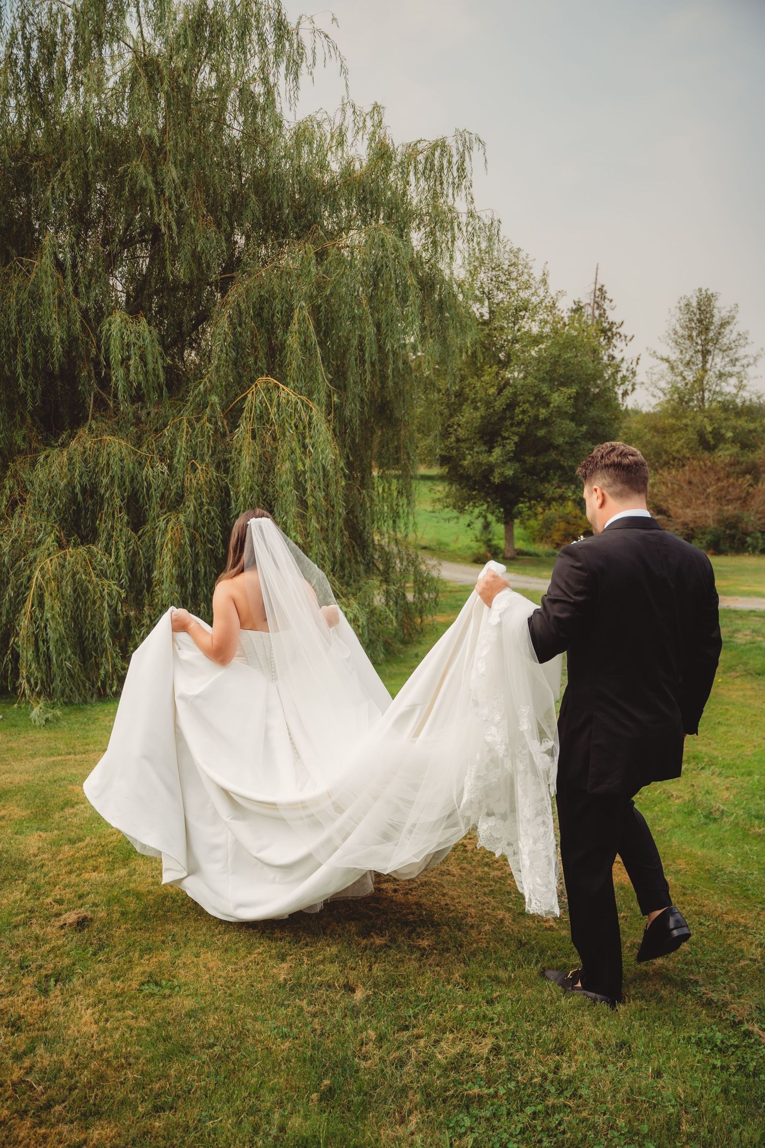 Groom in black suit helping carry bride's flowing white wedding dress train by a weeping willow.