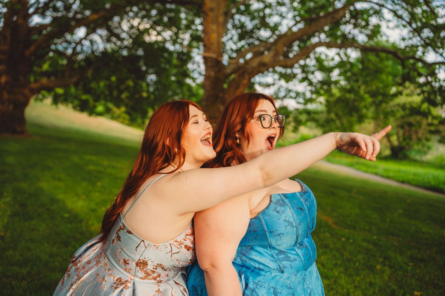 Two redhead women in floral dresses laughing and pointing outdoors under large green tree.