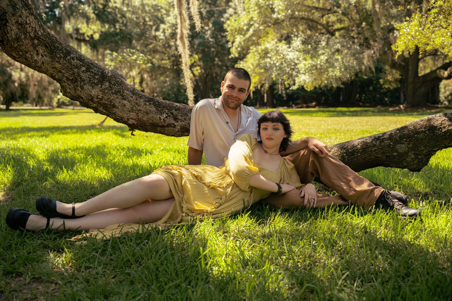 Couple relaxing under a tree on green grass, woman in yellow dress, man in beige shirt, sunny park setting.