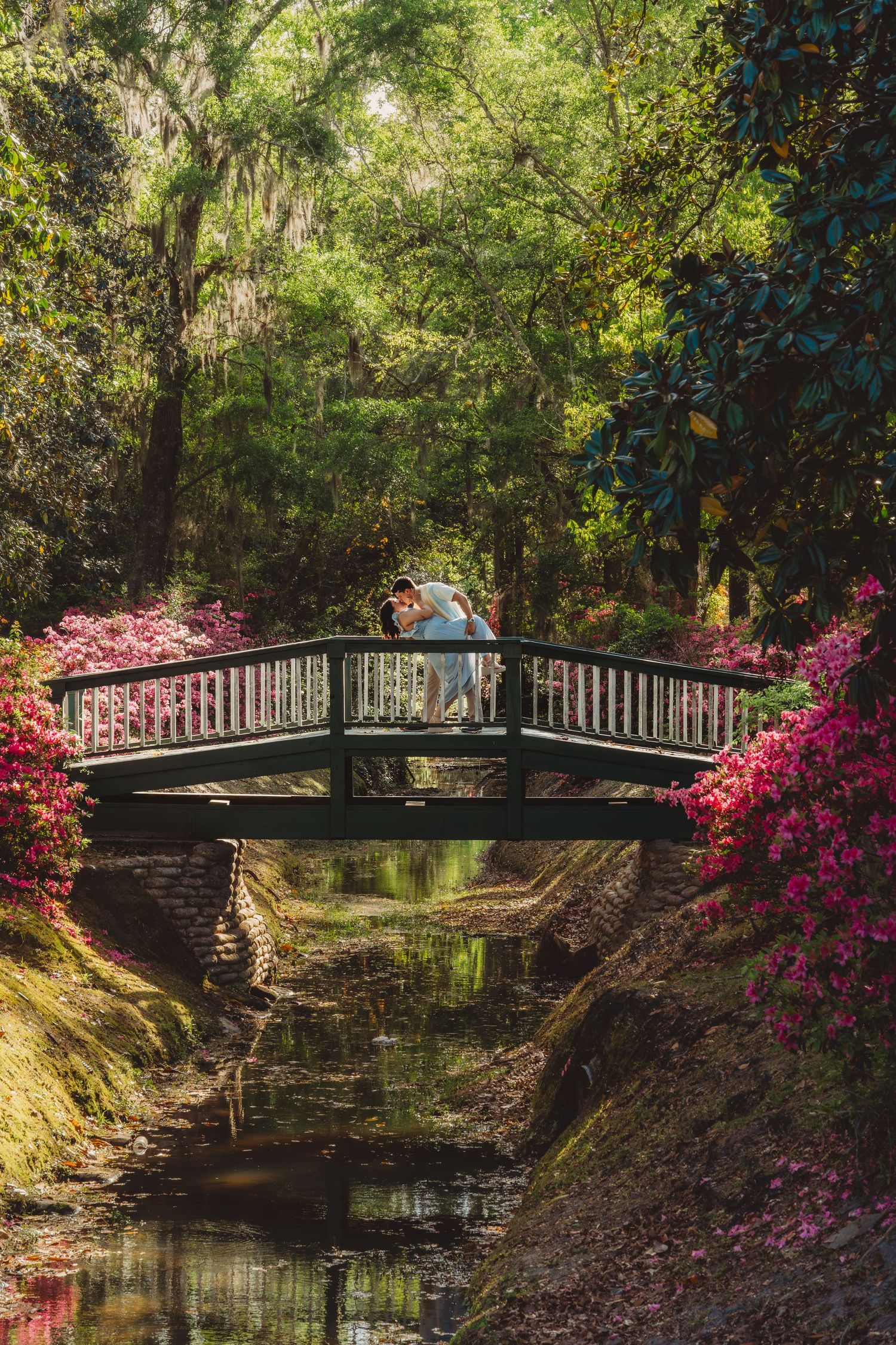 Couple kissing on a wooden bridge surrounded by blooming azaleas and lush green trees reflected in calm water.