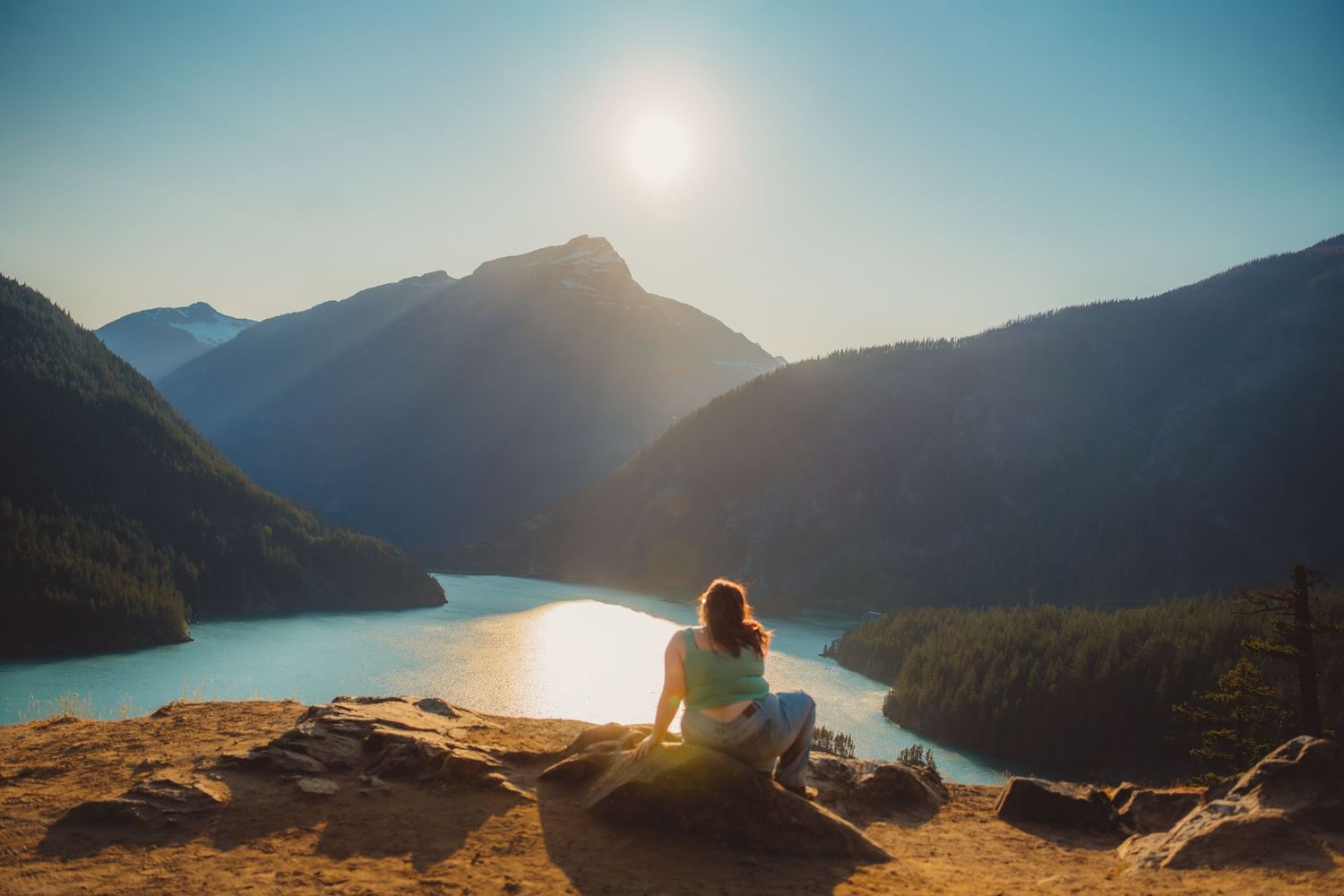Woman sitting on rocky overlook gazing at a serene mountain lake surrounded by peaks bathed in golden sunlight.