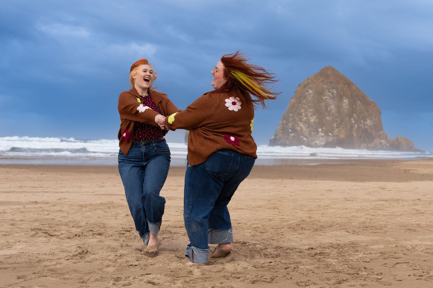 Two women in matching brown sweaters and jeans laugh and dance on a sandy beach with Haystack Rock in the background.