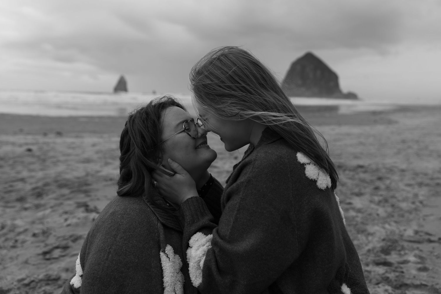 Two women share a tender kiss on a dramatic black and white beach with Haystack Rock in the background.