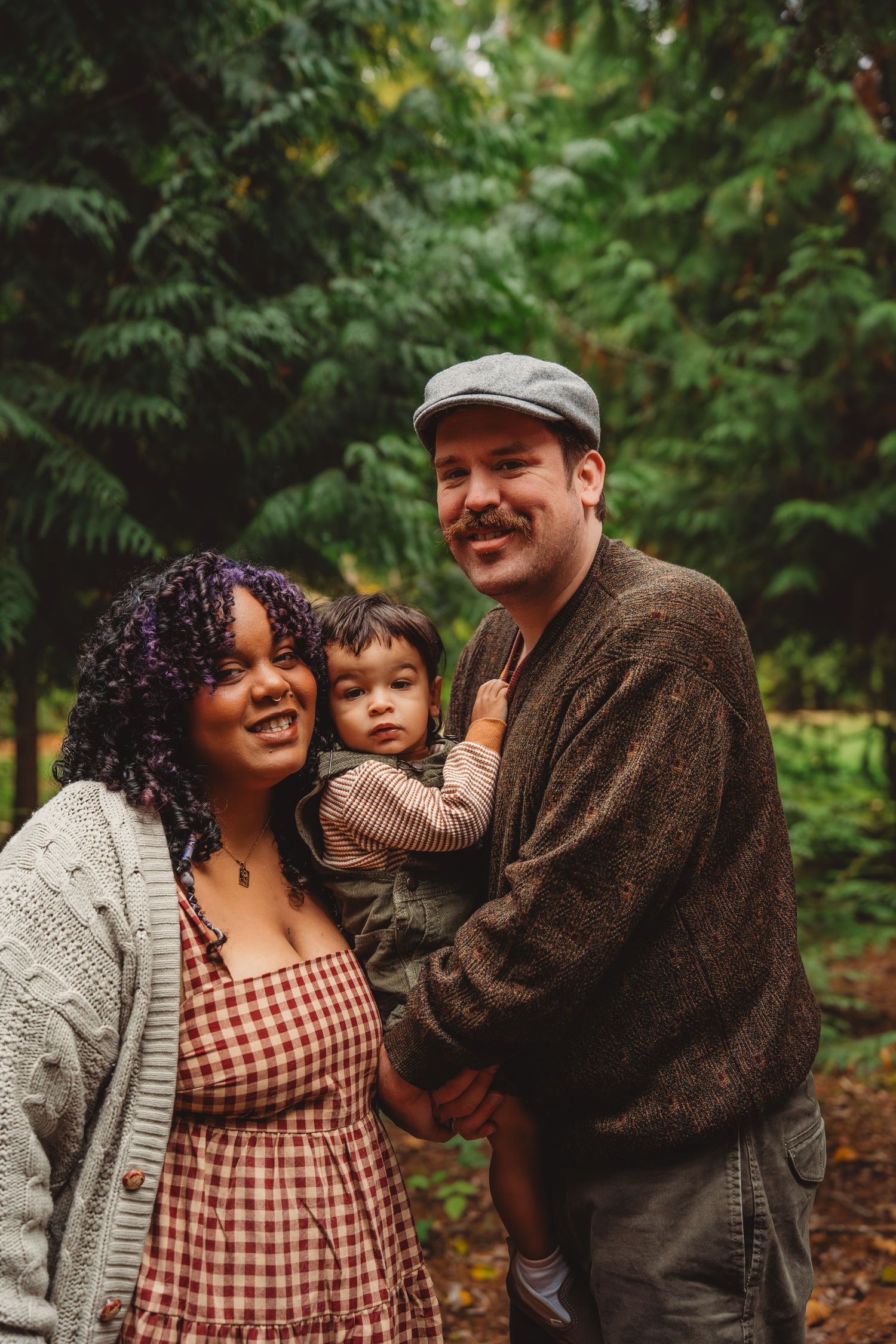 A smiling interracial family with a young toddler poses outdoors among lush green trees in a park.
