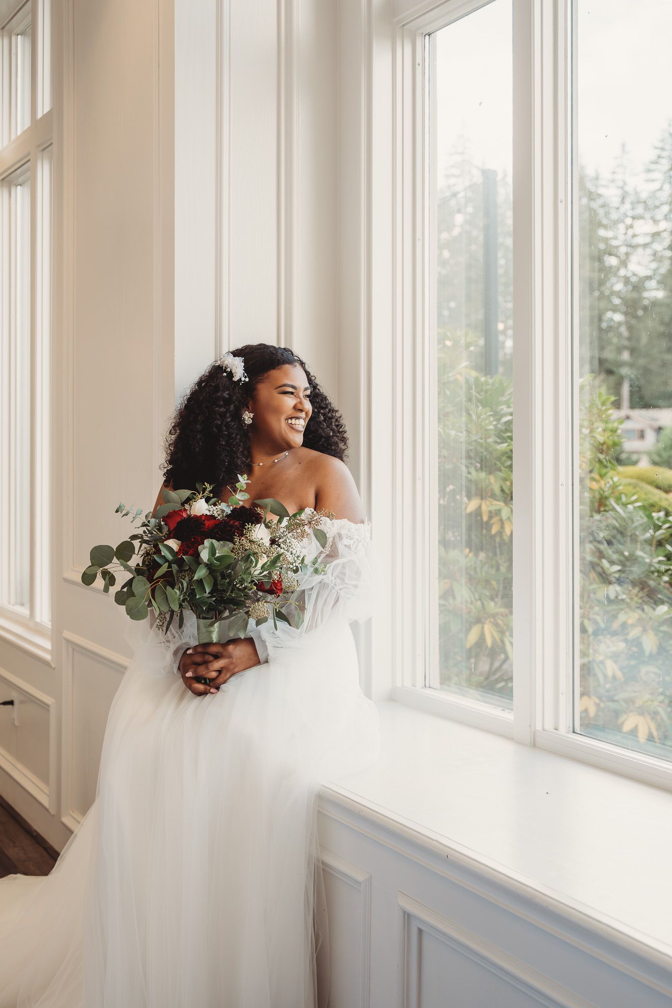 Smiling bride in an off-shoulder tulle gown holds a red and white bouquet while standing by bright windows.