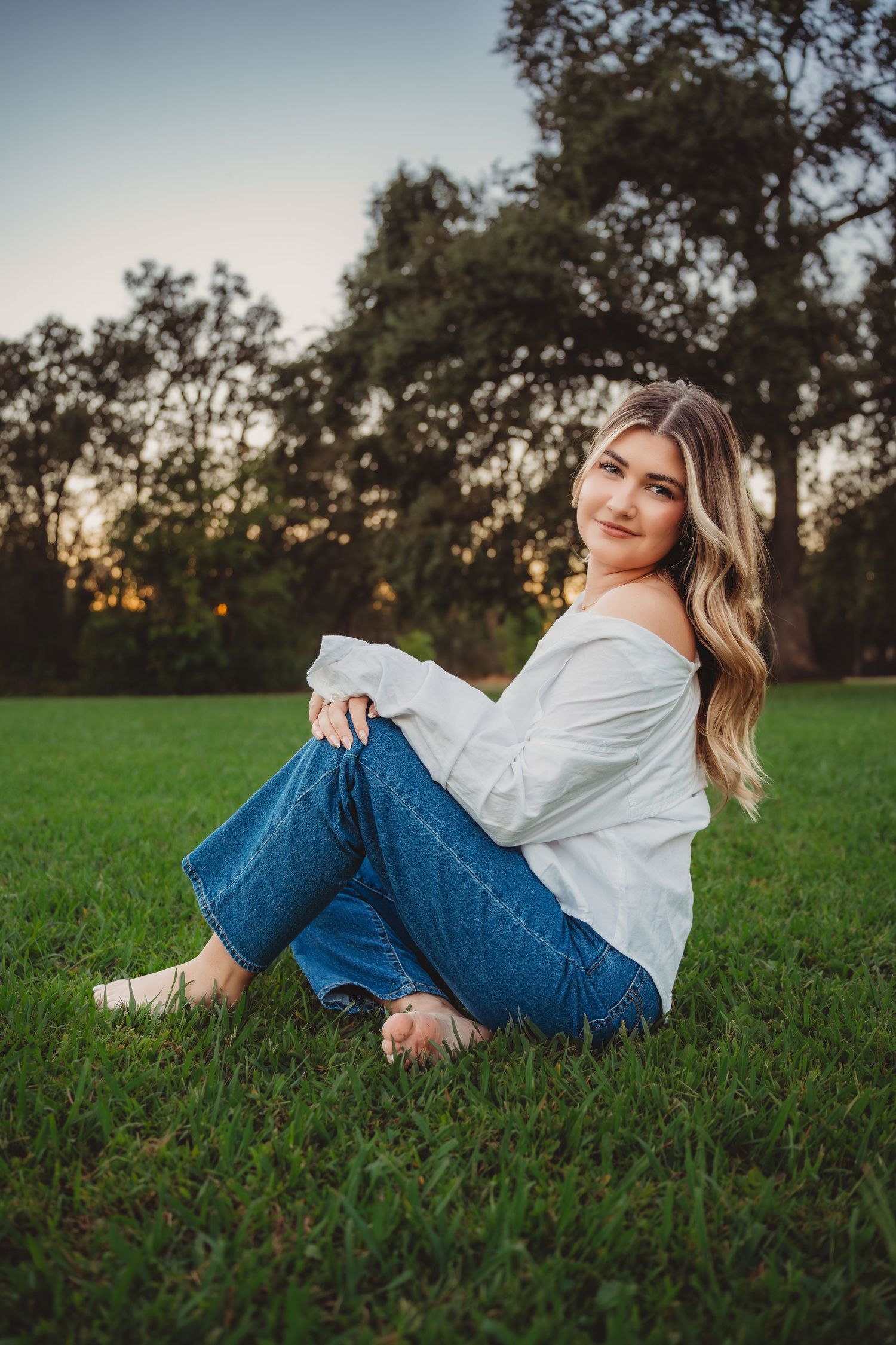 Blonde woman in white top and jeans sitting on grass at golden hour.