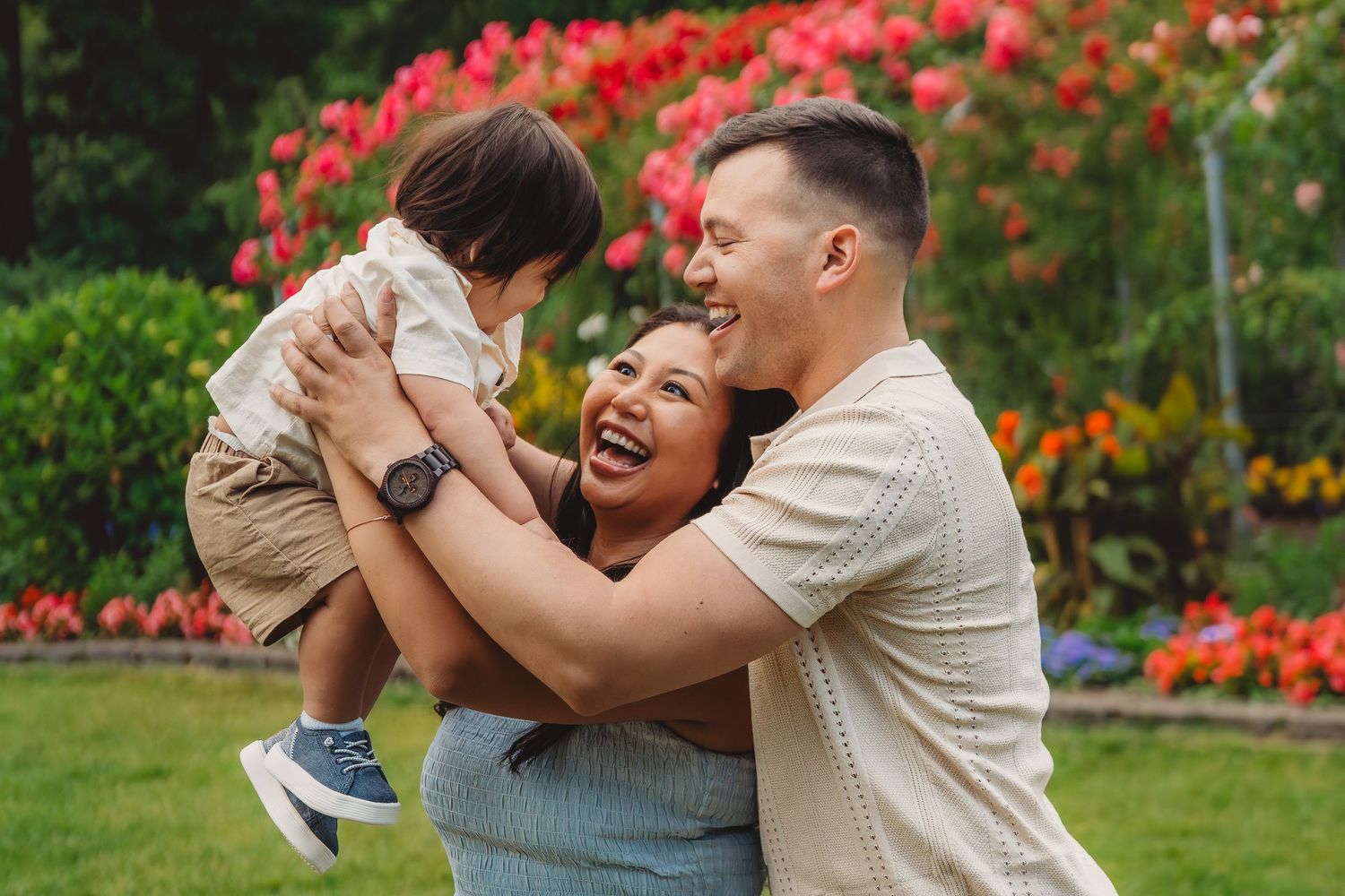 Happy family with parents laughing and lifting their baby boy outdoors near colorful flower garden.