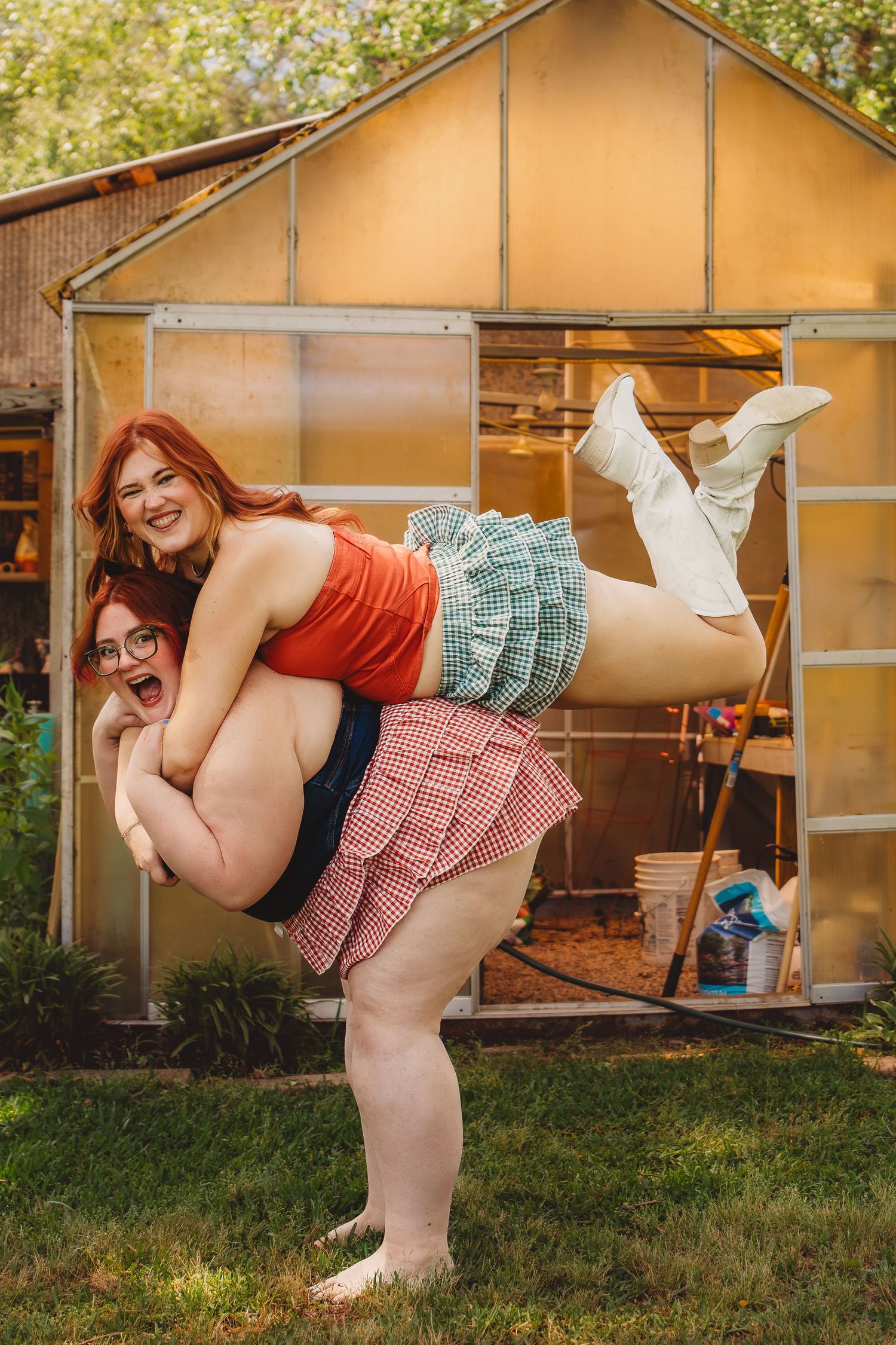 Two redhead women laughing outdoors, one giving the other a piggyback ride in front of a greenhouse.