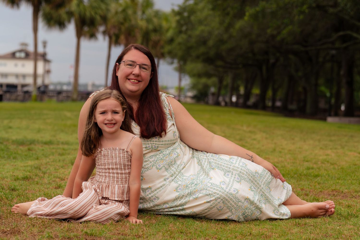 A woman and young girl smile together on green grass in a park, wearing summer dresses on a cloudy day.