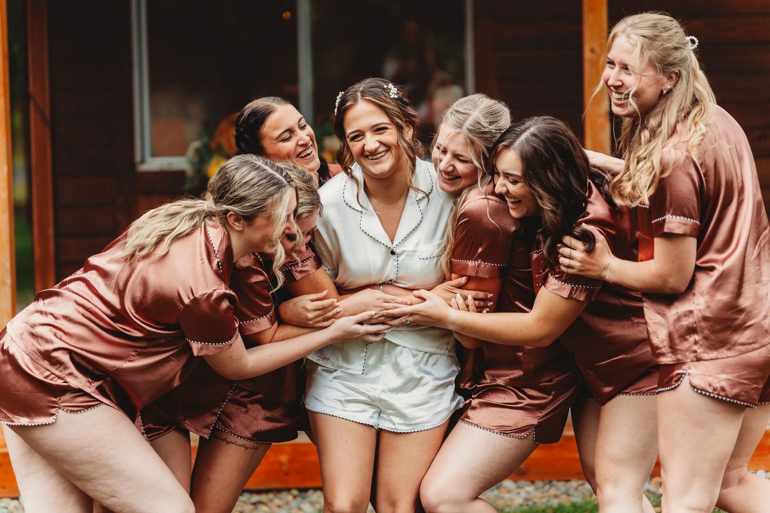 Bride in white pajamas surrounded by bridesmaids in matching brown satin sets, laughing together before a wedding.