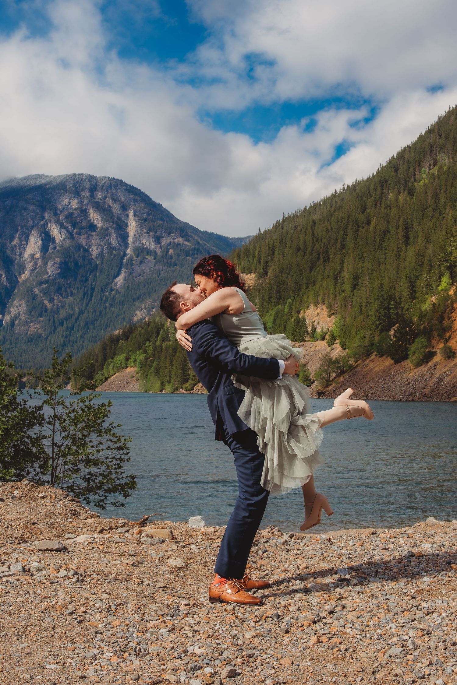 Couple sharing a romantic kiss by a mountain lake, groom lifting bride in wedding attire with scenic forest backdrop.